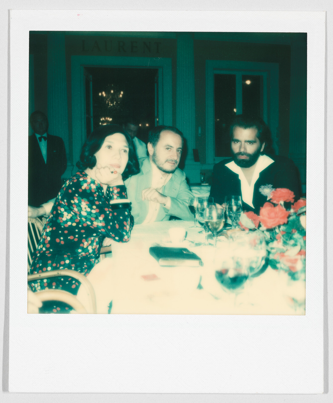 Three people sit at a dinner table with drinks and flowers, under a sign that reads "LAURENT."