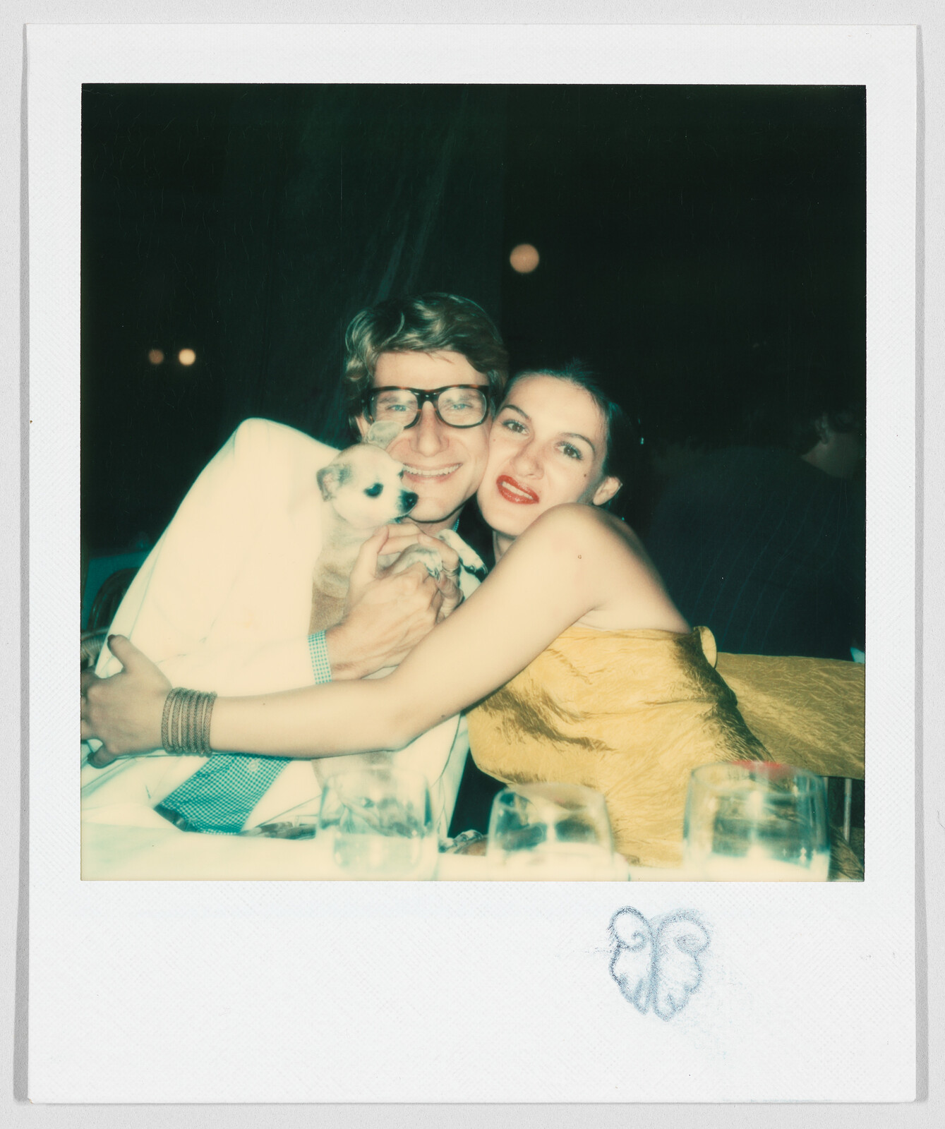 Two people smiling and hugging at a table, holding a small dog between them, with glasses in front.