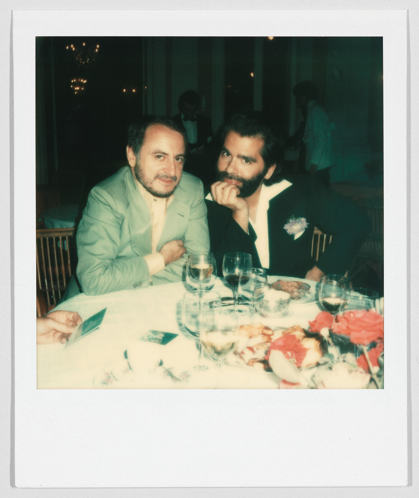Two men sit closely at a dinner table with drinks, flowers, and food, looking toward the camera.