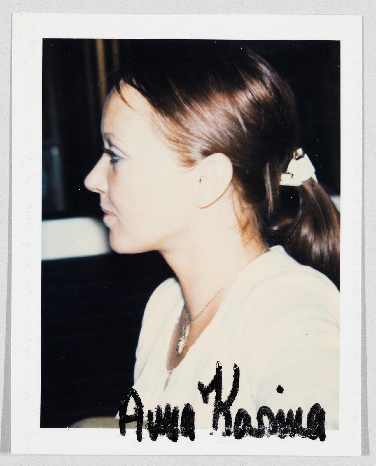 Woman with brown hair in a ponytail, wearing a white top and necklace. "Anna Karina" is written below.