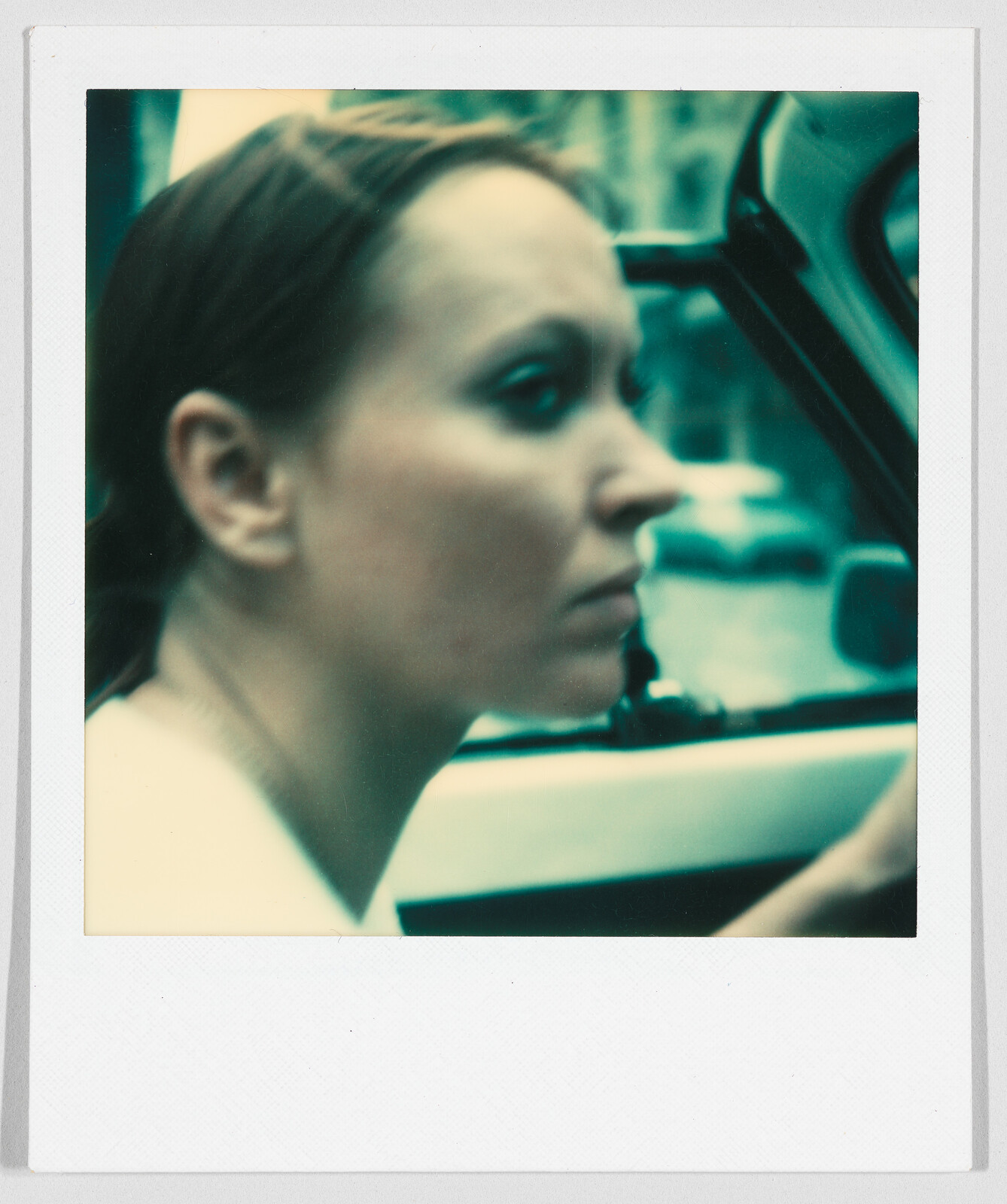 Woman with brown hair driving a car, looking forward with a serious expression. Background is blurry.