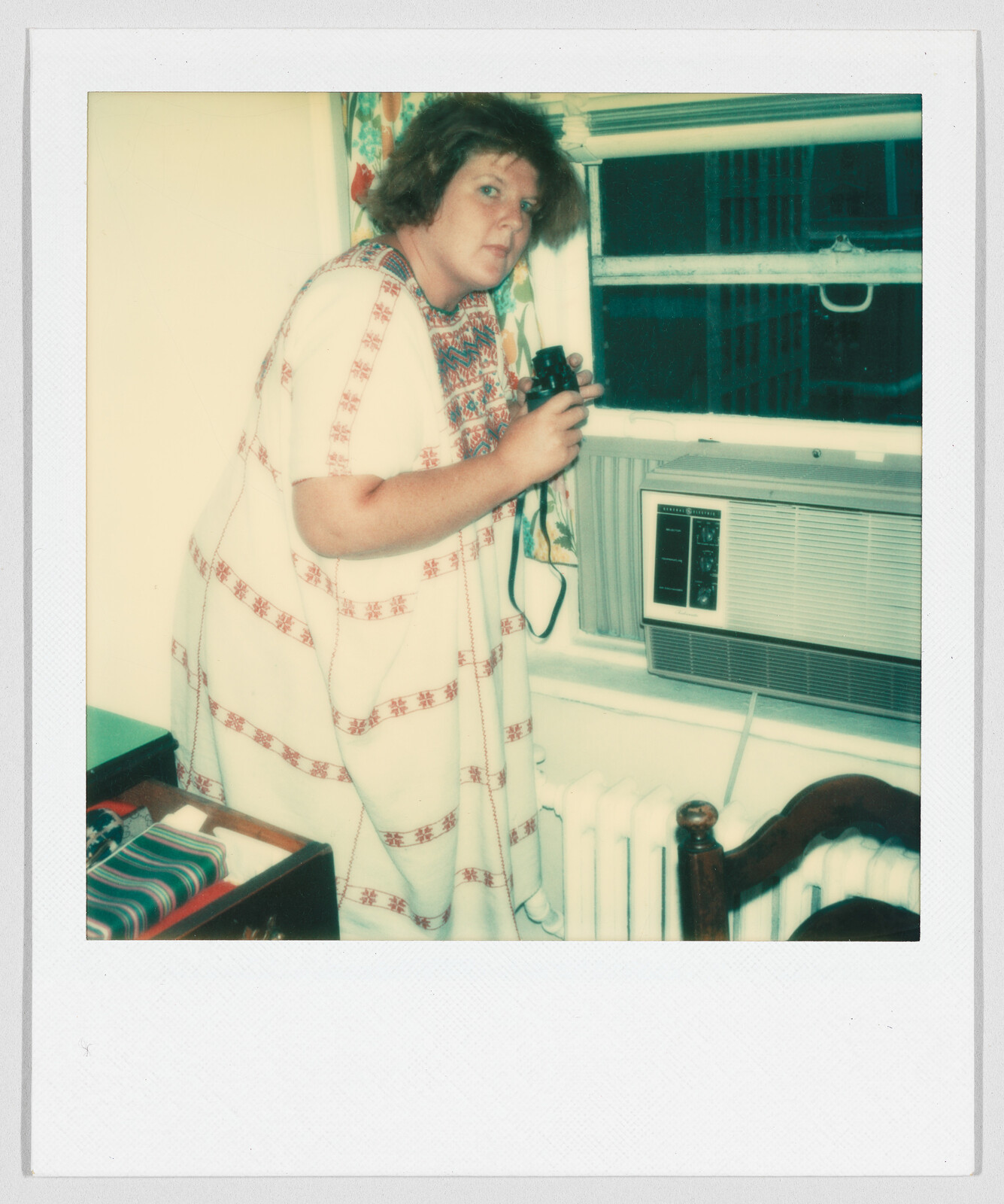 Woman in a patterned dress holding binoculars, looking out of a window next to an air conditioner.