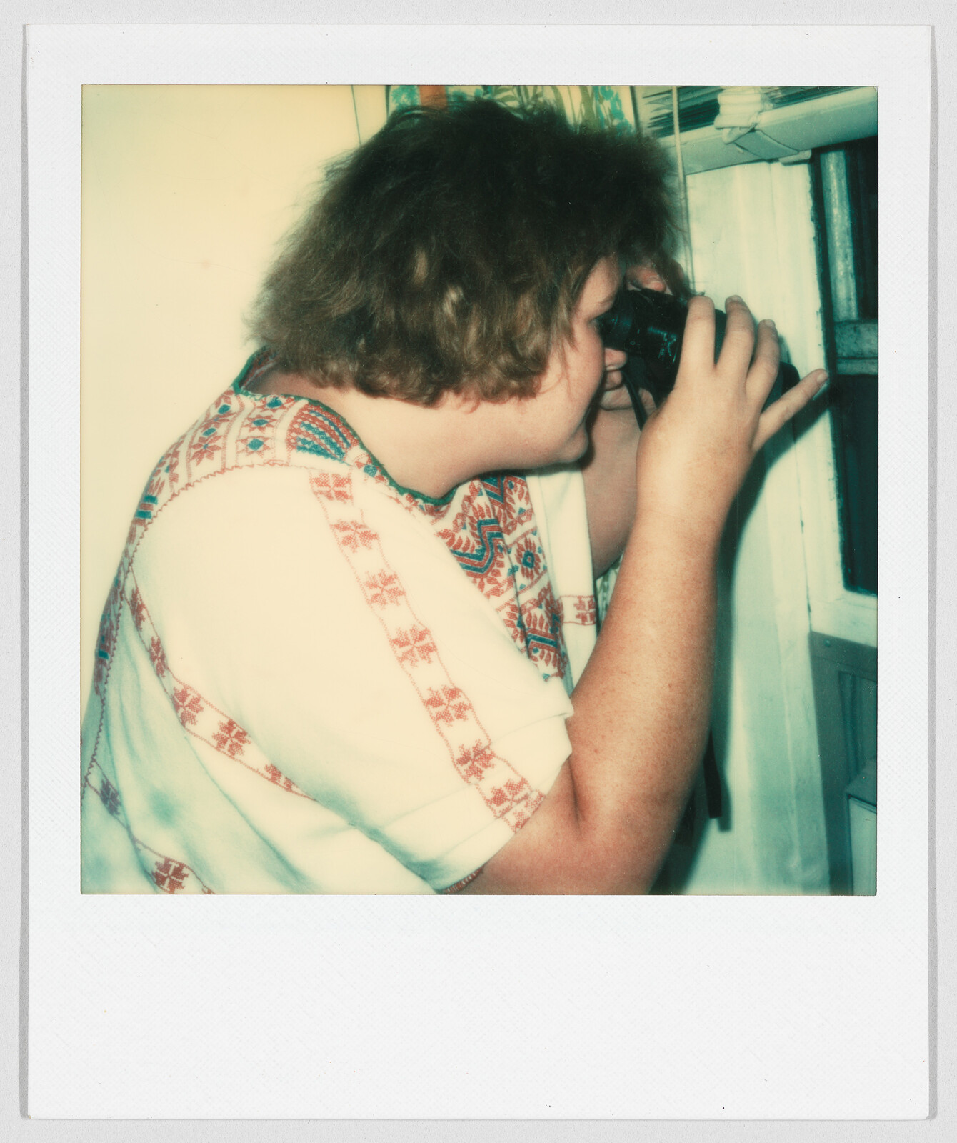 Person with curly hair looks through binoculars while standing next to a window inside a room.