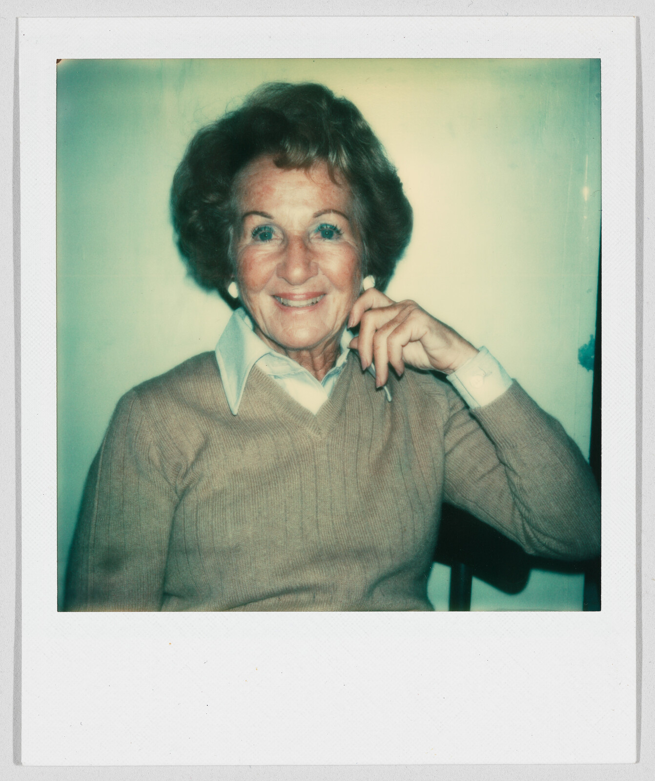 Older woman with short hair and a tan sweater sits and smiles, resting her hand on her chin.