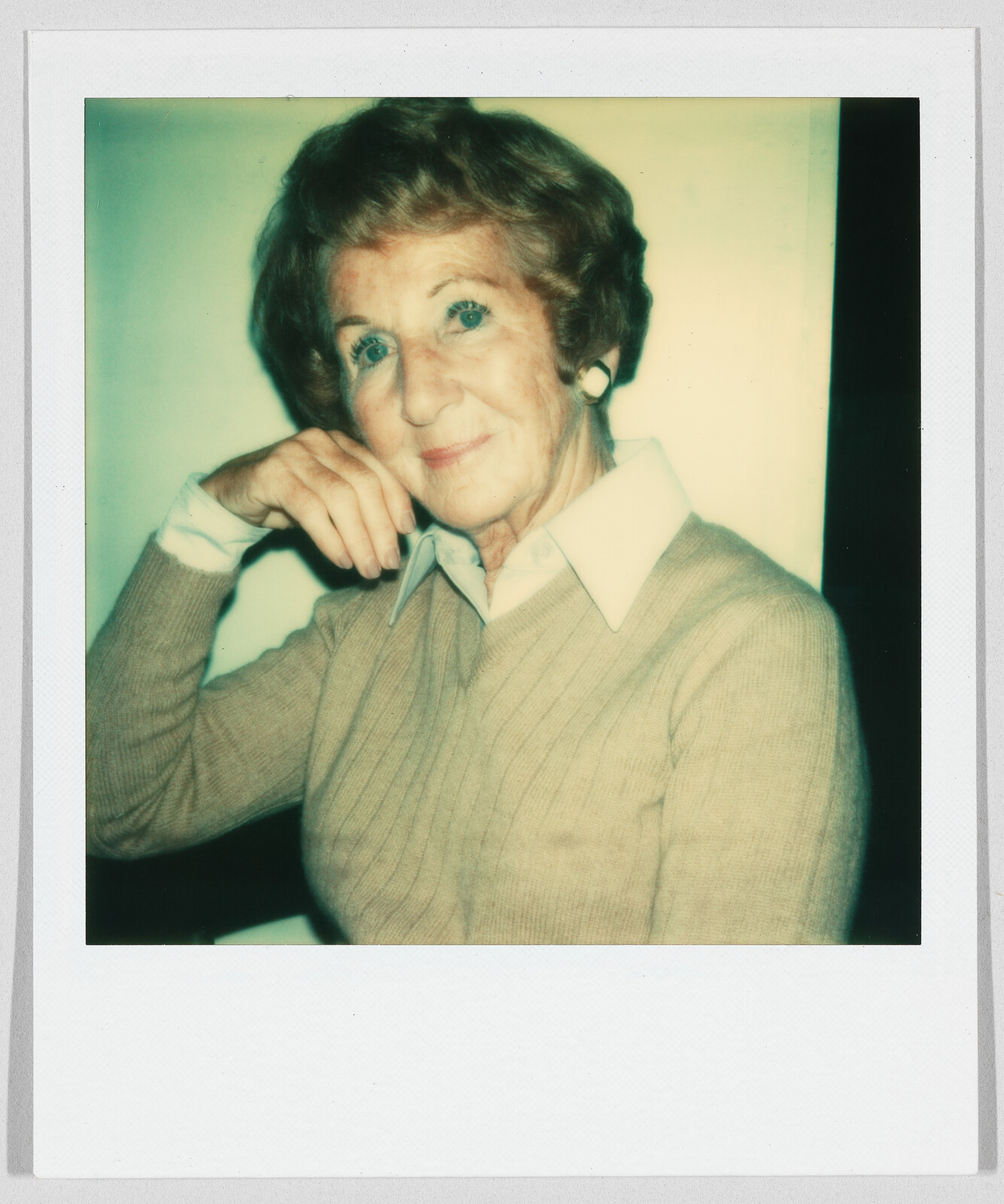 Older woman with short hair and earrings sits, resting her chin on her hand, looking at the camera.