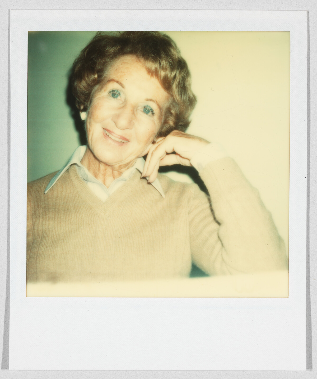Older woman with short hair smiles and rests her head on her hand, wearing a tan sweater and collared shirt.