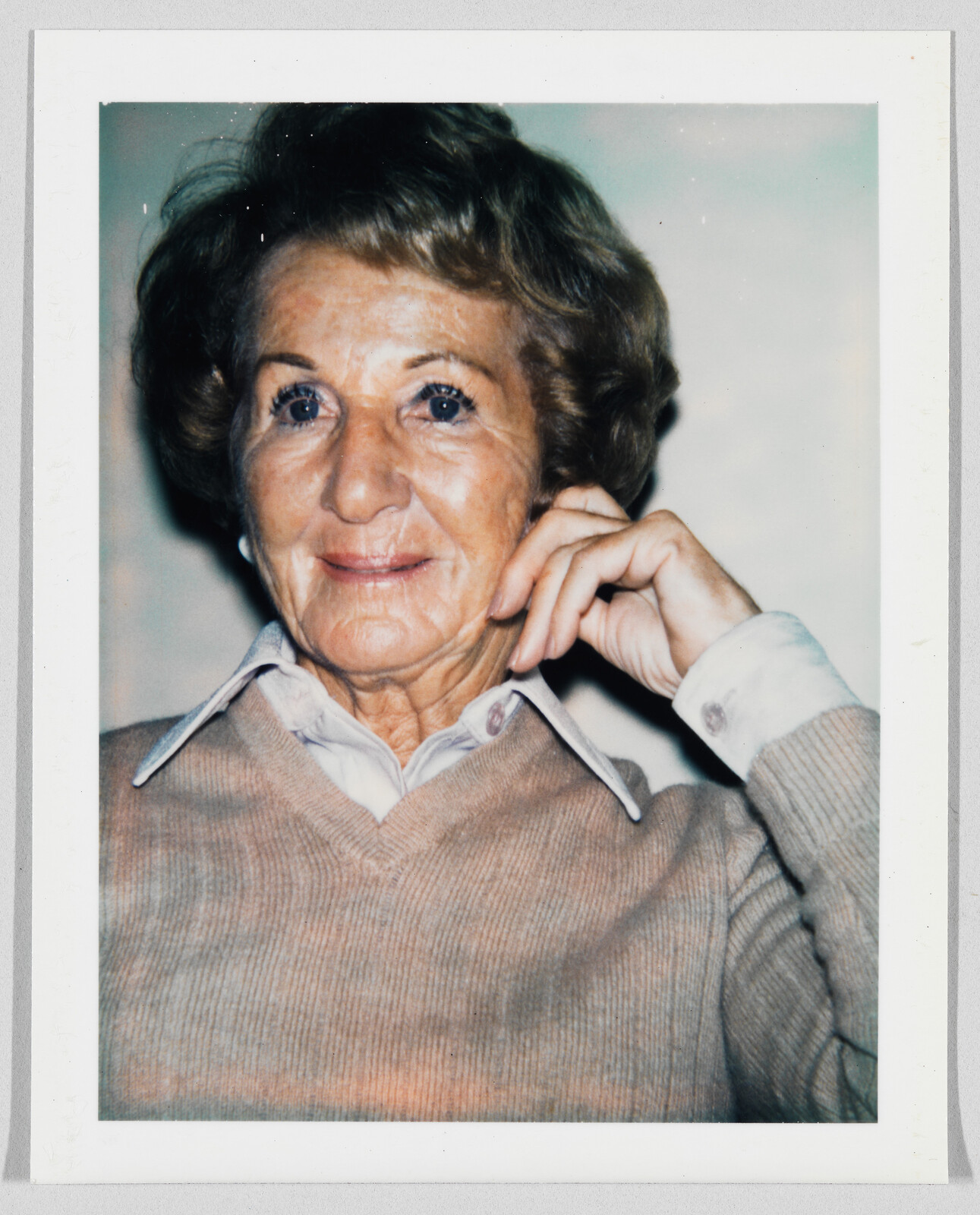 Older woman with short hair wearing a beige sweater and collared shirt, resting her hand on her face.