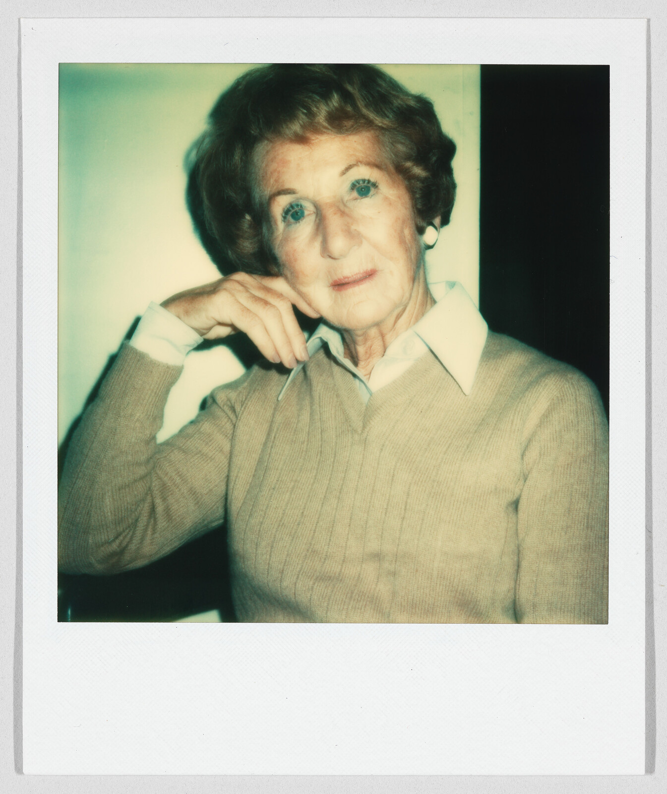 Older woman with short hair wearing a tan sweater and white collared shirt, resting her hand on her face.