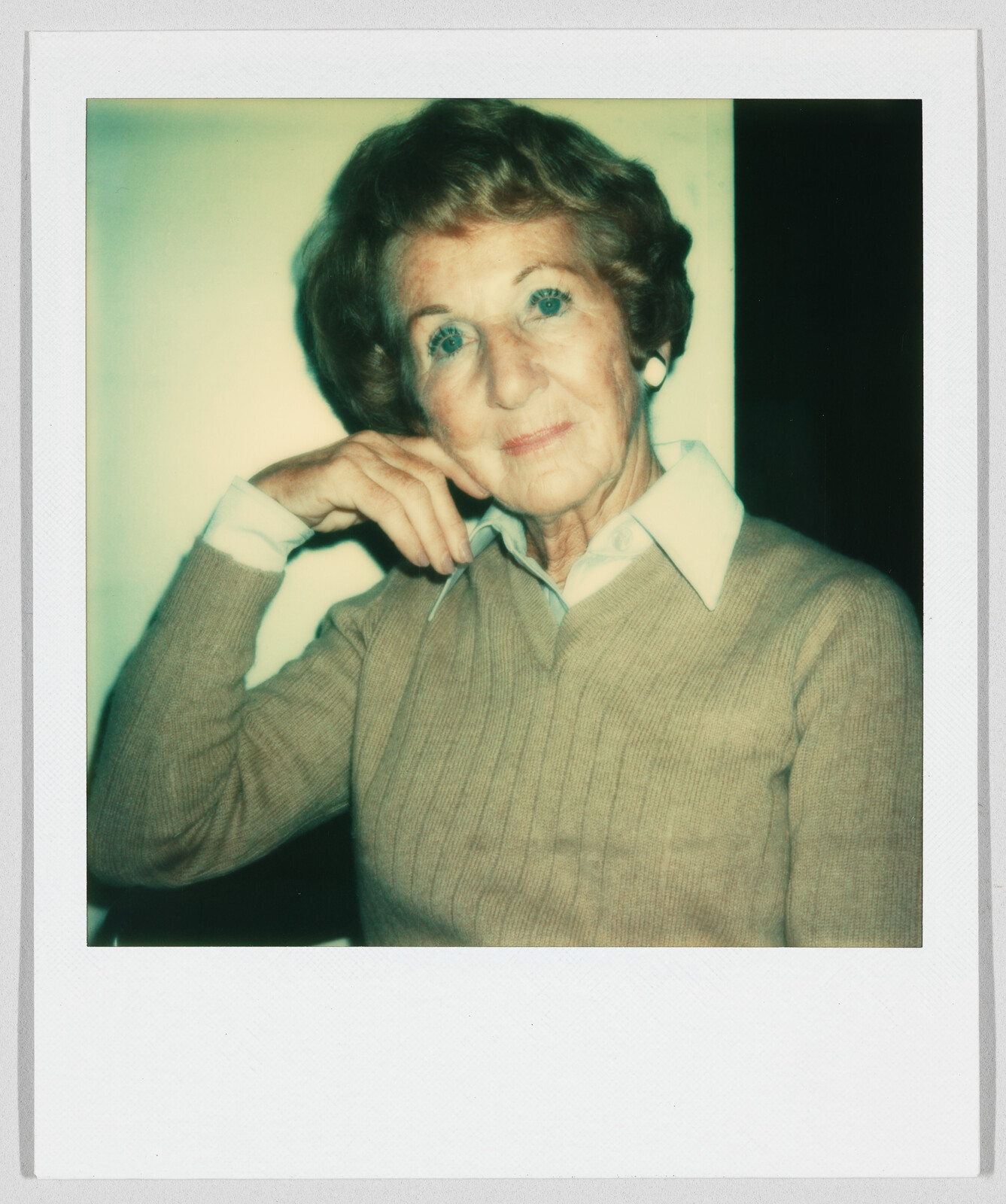 Older woman with short hair and a tan sweater sits, resting her head on her hand, looking forward.