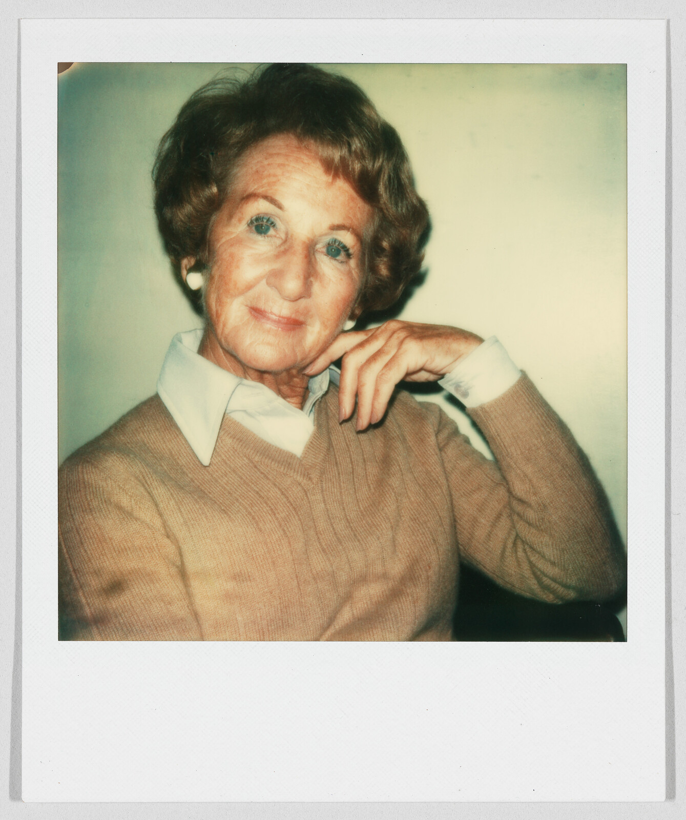 Older woman with short hair and a tan sweater sits, resting her chin on her hand, smiling gently.