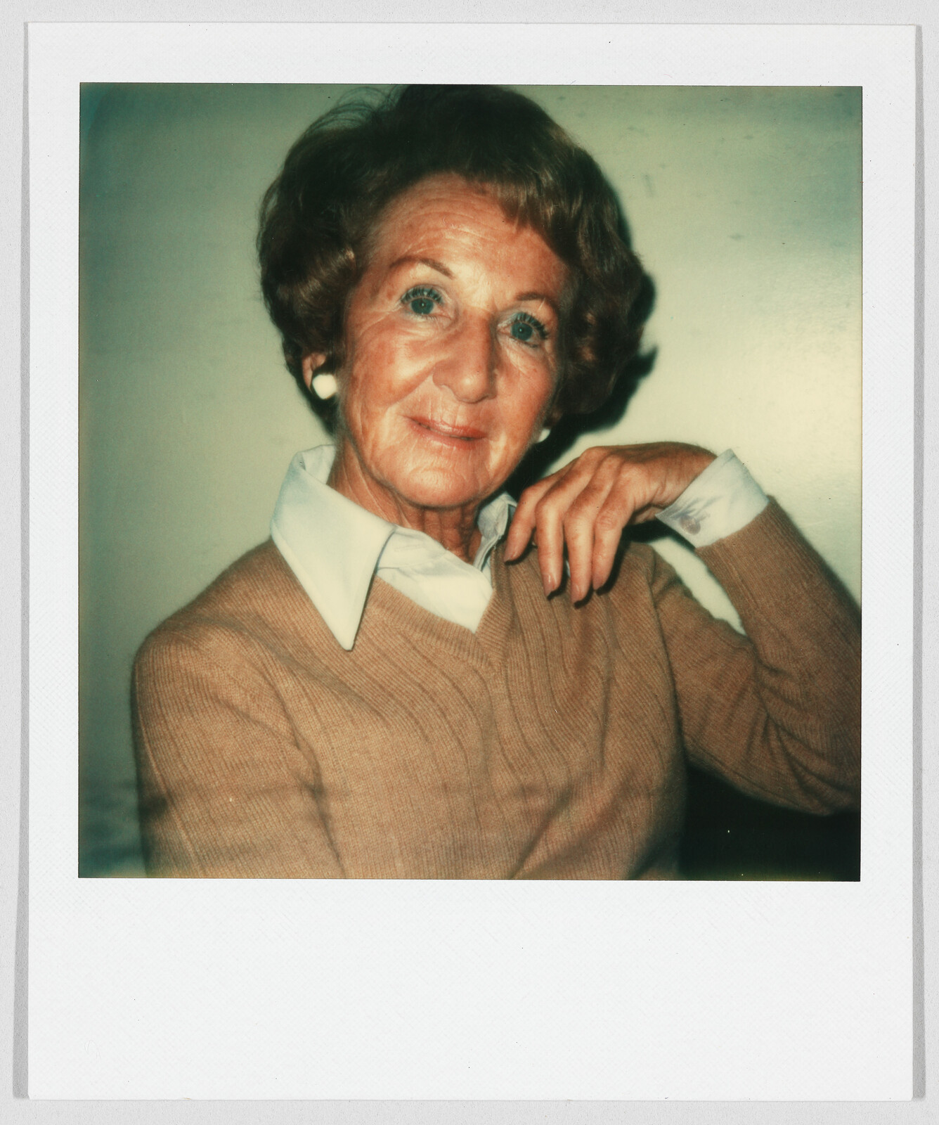 Older woman with short brown hair, wearing a tan sweater and white collared shirt, sitting and smiling gently.