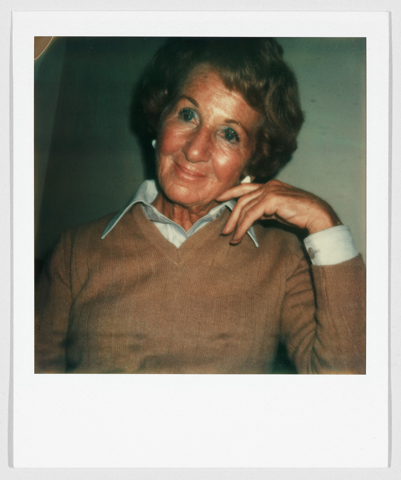 Older woman with short hair wearing a tan sweater and collared shirt, smiling and resting her hand on her face.