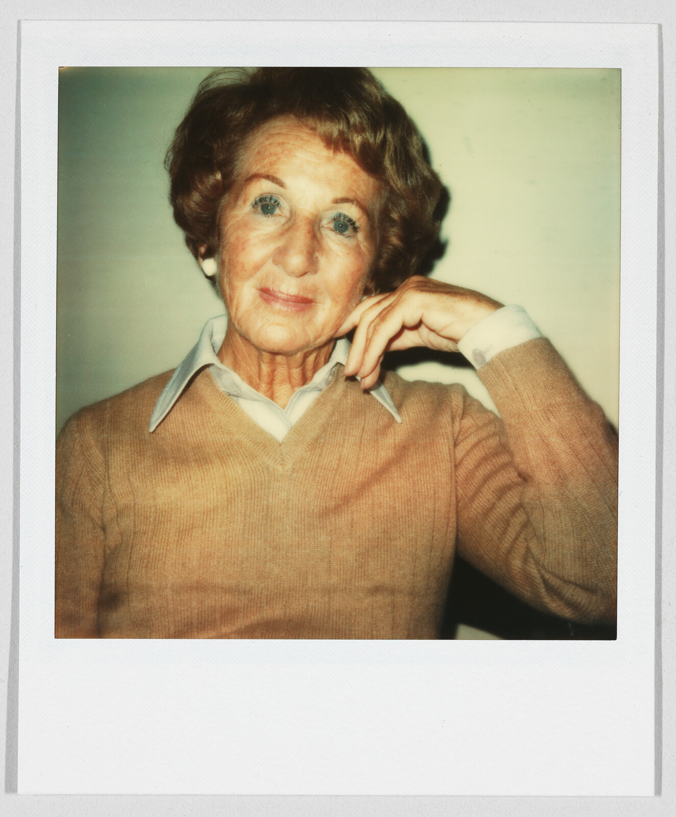 Older woman with short hair wearing a tan sweater and collared shirt, resting her hand on her chin.