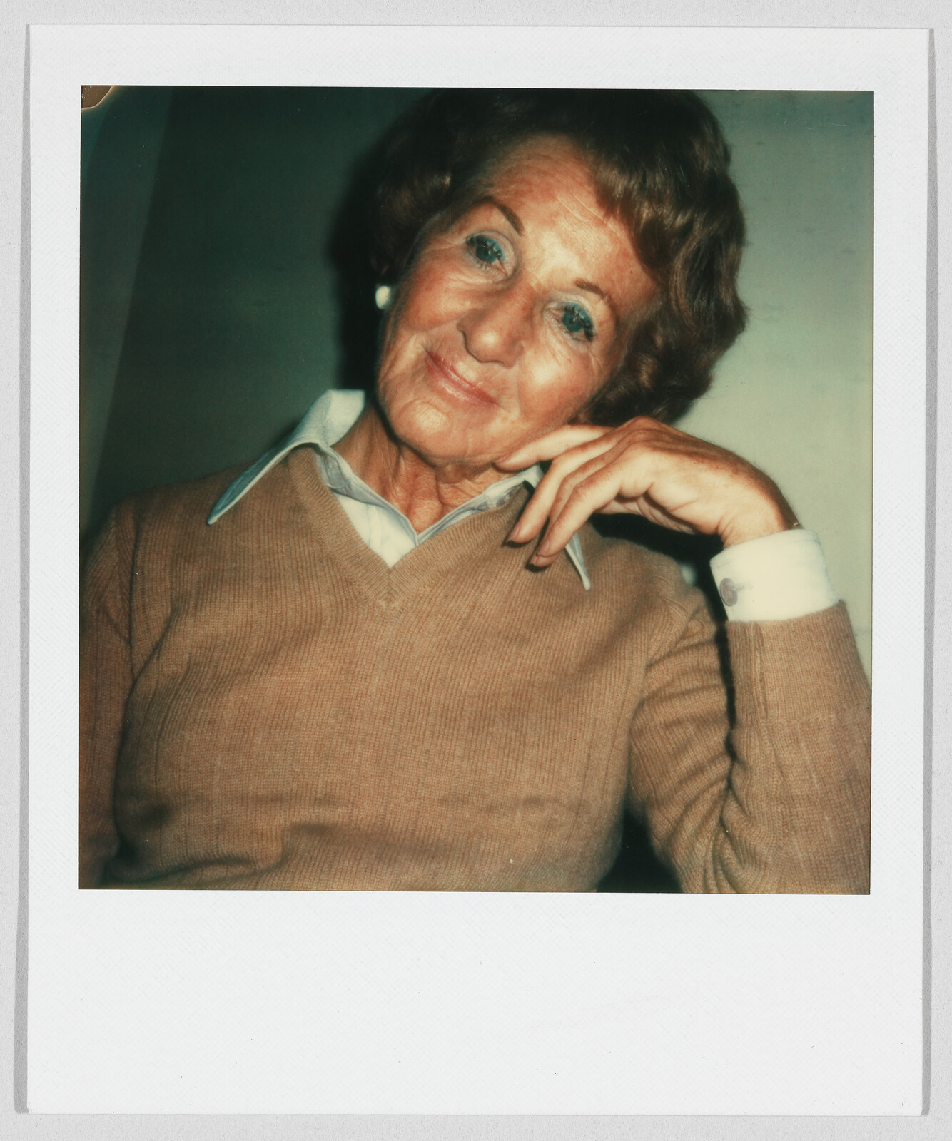 Older woman with short hair and a tan sweater sits, resting her head on her hand, smiling gently.