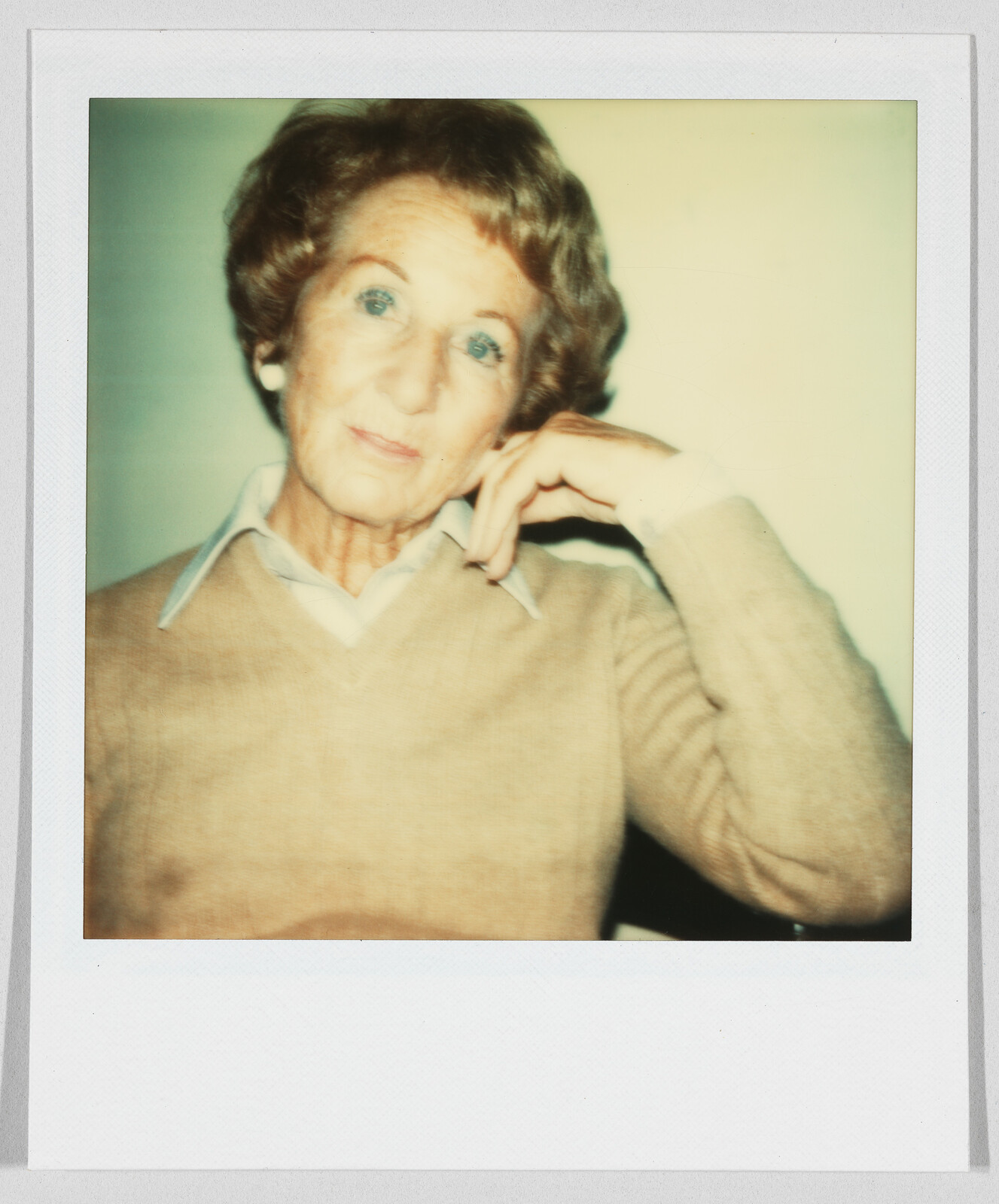 Older woman with short hair wearing a beige sweater, resting her head on her hand and looking forward.