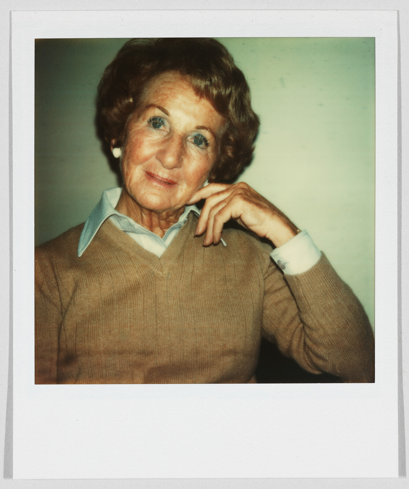 Older woman with short hair wearing a tan sweater and collared shirt, resting her hand on her face.