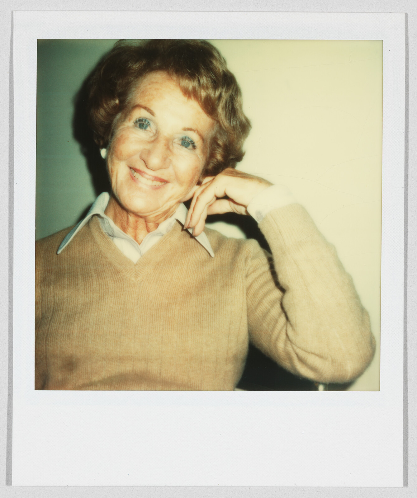 Older woman with short hair smiles warmly, resting her head on her hand, wearing a tan sweater.