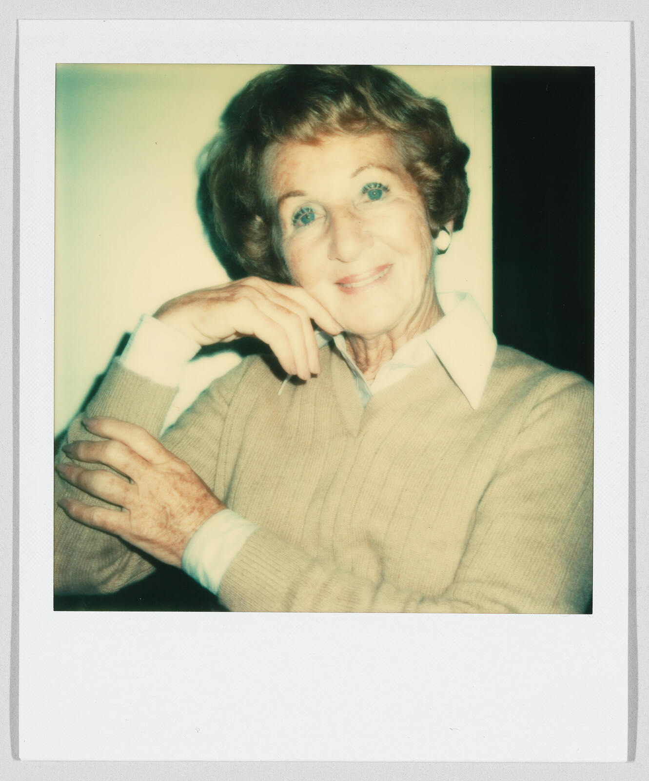 An older woman with short hair smiles gently, resting her chin on her hand and wearing a beige sweater.