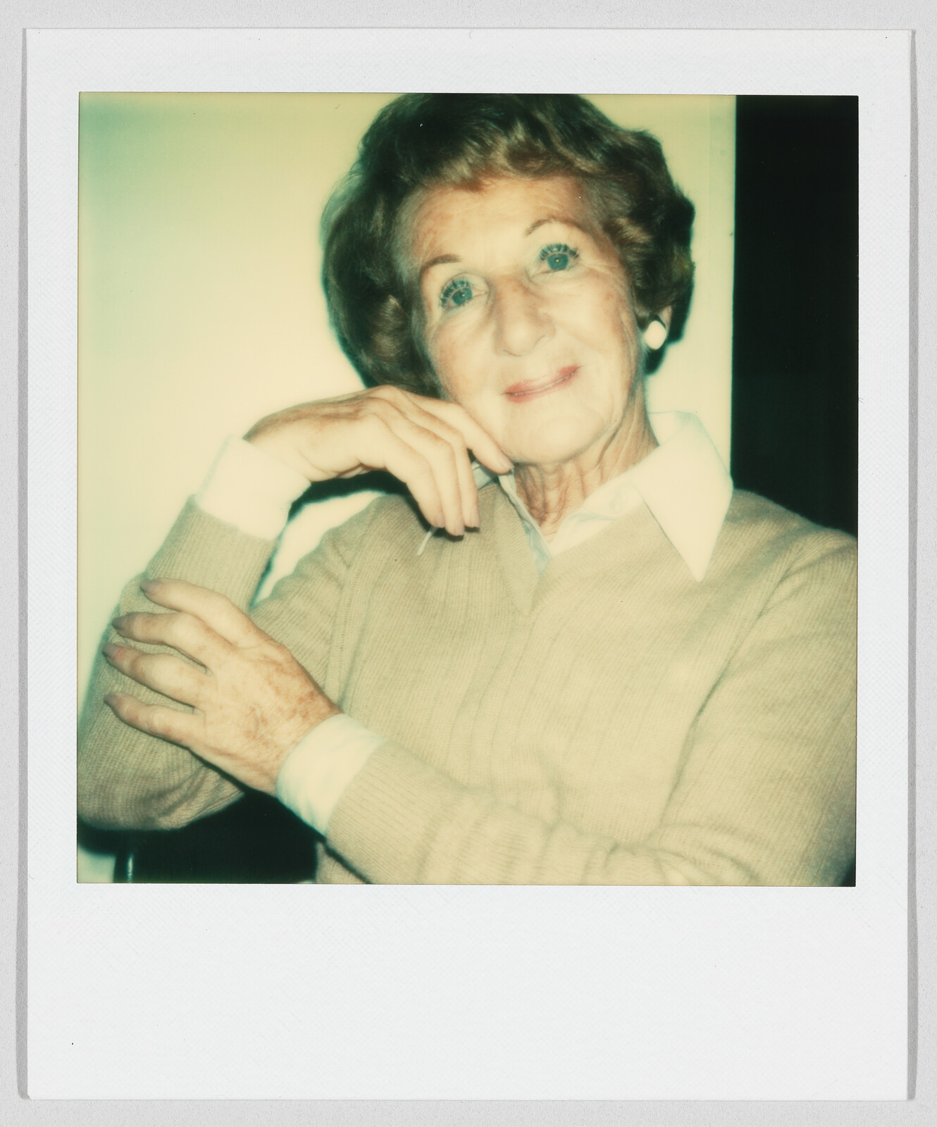 Older woman with short hair and a beige sweater sits, resting her chin on her hand, smiling gently.
