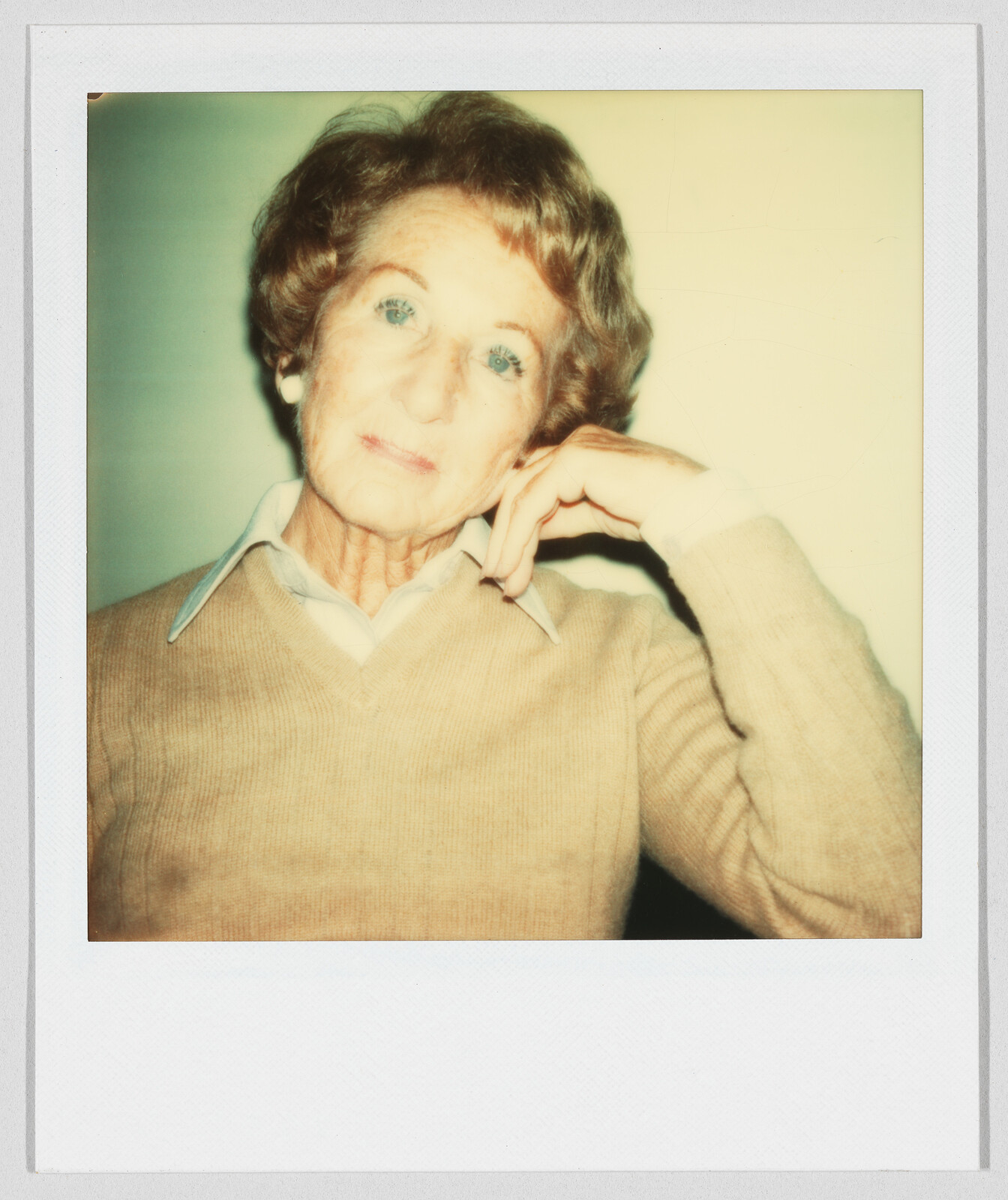 Older woman with short hair wearing a tan sweater, resting her head on her hand, looking at camera.