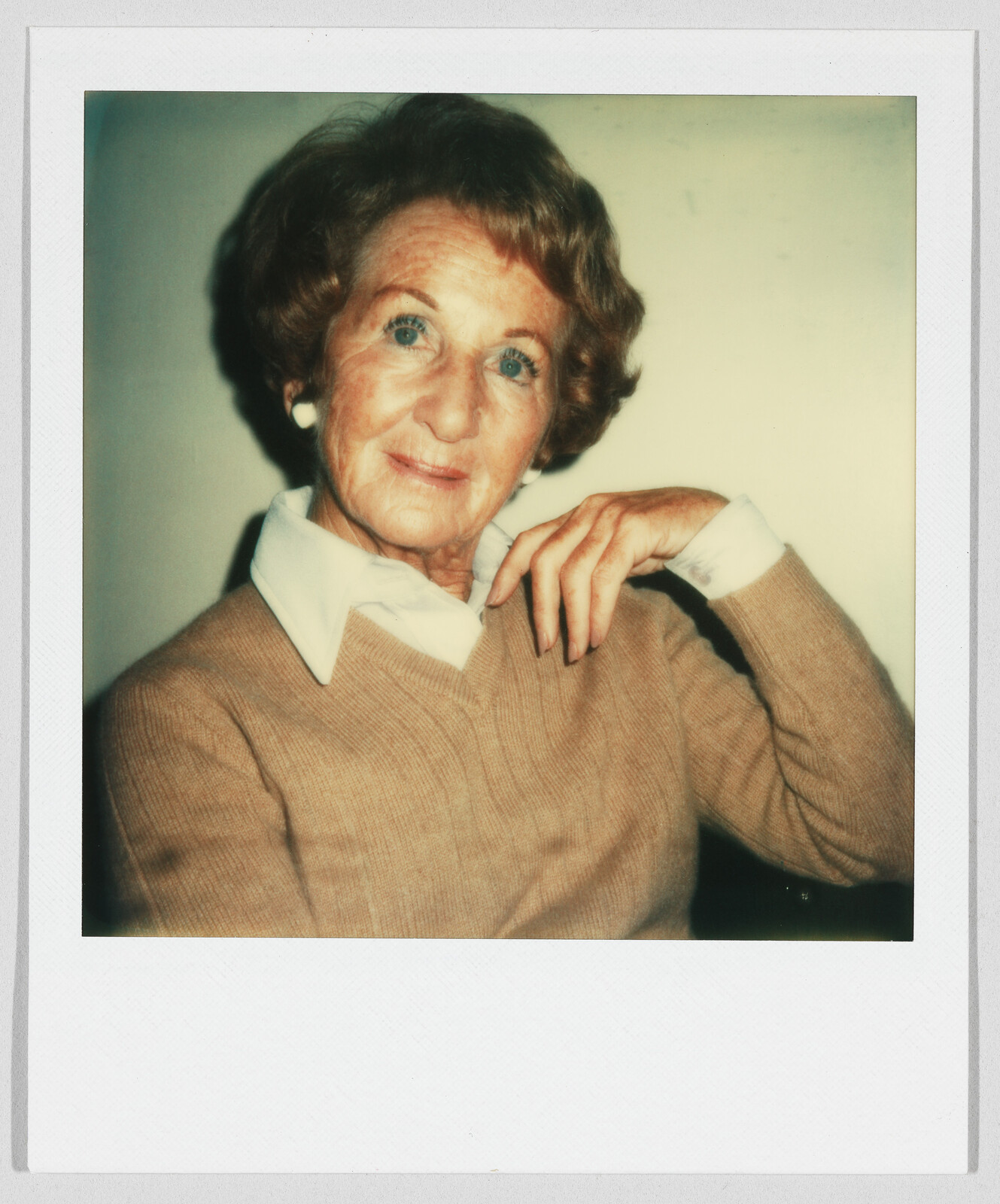 Older woman with short hair and a tan sweater sits and looks at the camera, hand near her face.