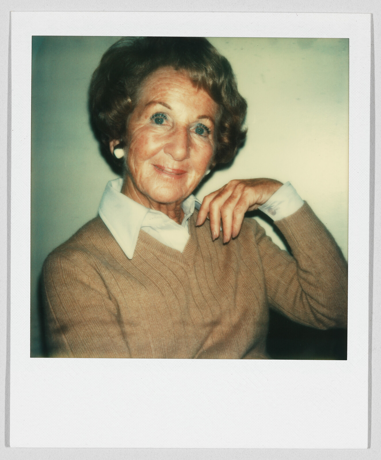 Older woman with short hair, wearing a tan sweater and white collared shirt, smiling and posing indoors.