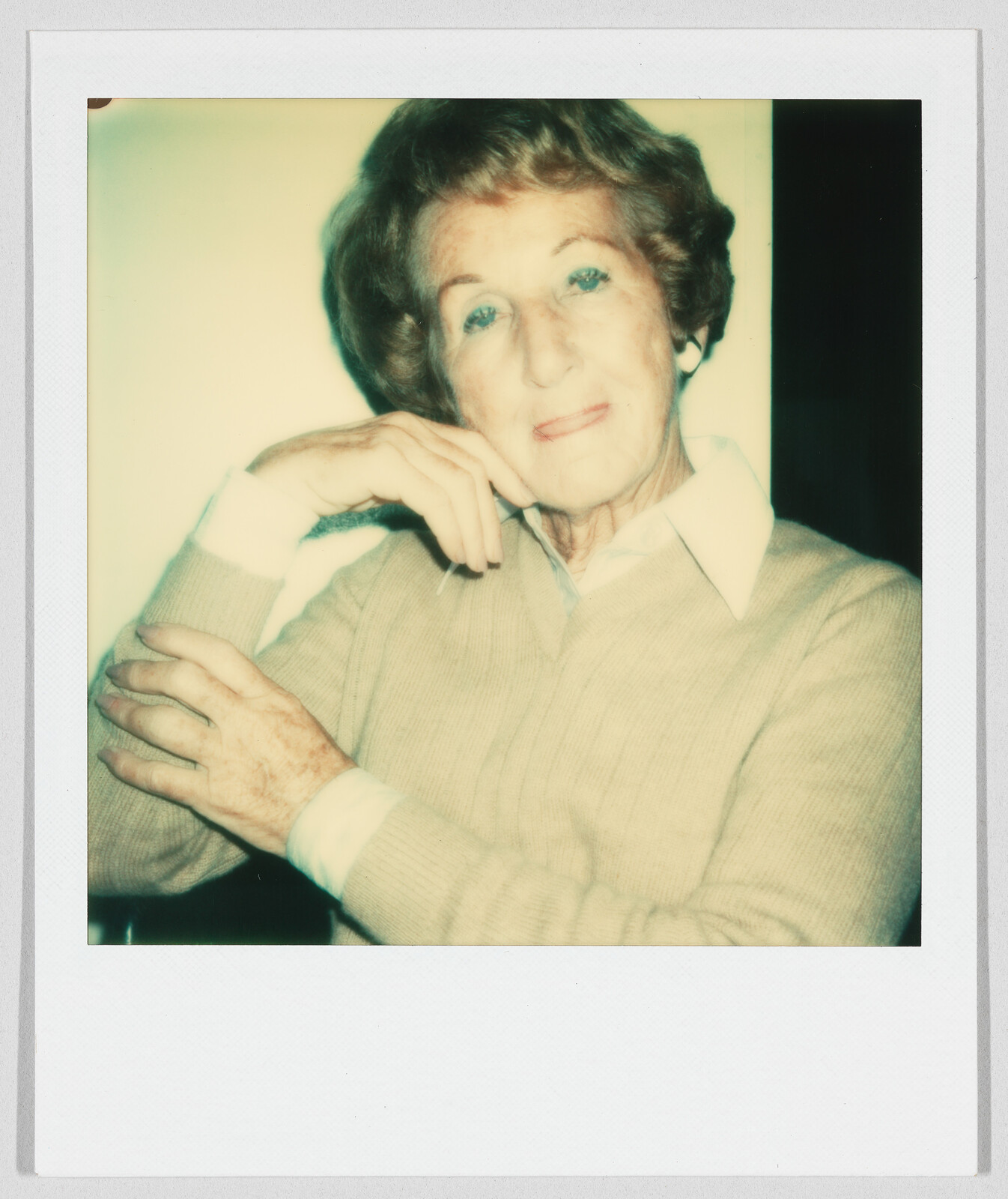An older woman with short hair and a beige sweater sits with one arm resting on the other.