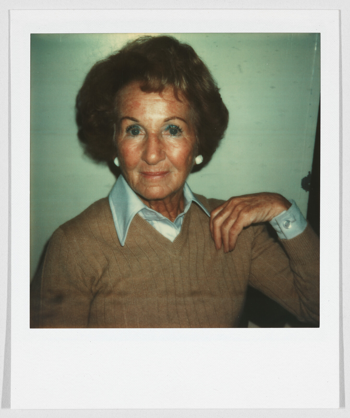Older woman with short brown hair, wearing a tan sweater and blue collared shirt, sitting and looking forward.