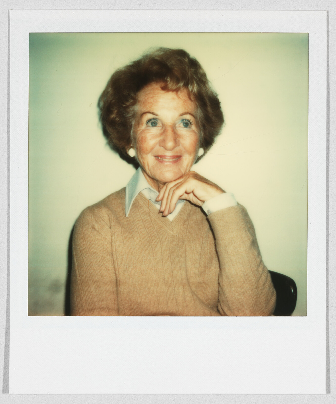 Older woman with short hair and a tan sweater smiles, resting her chin on her hand.