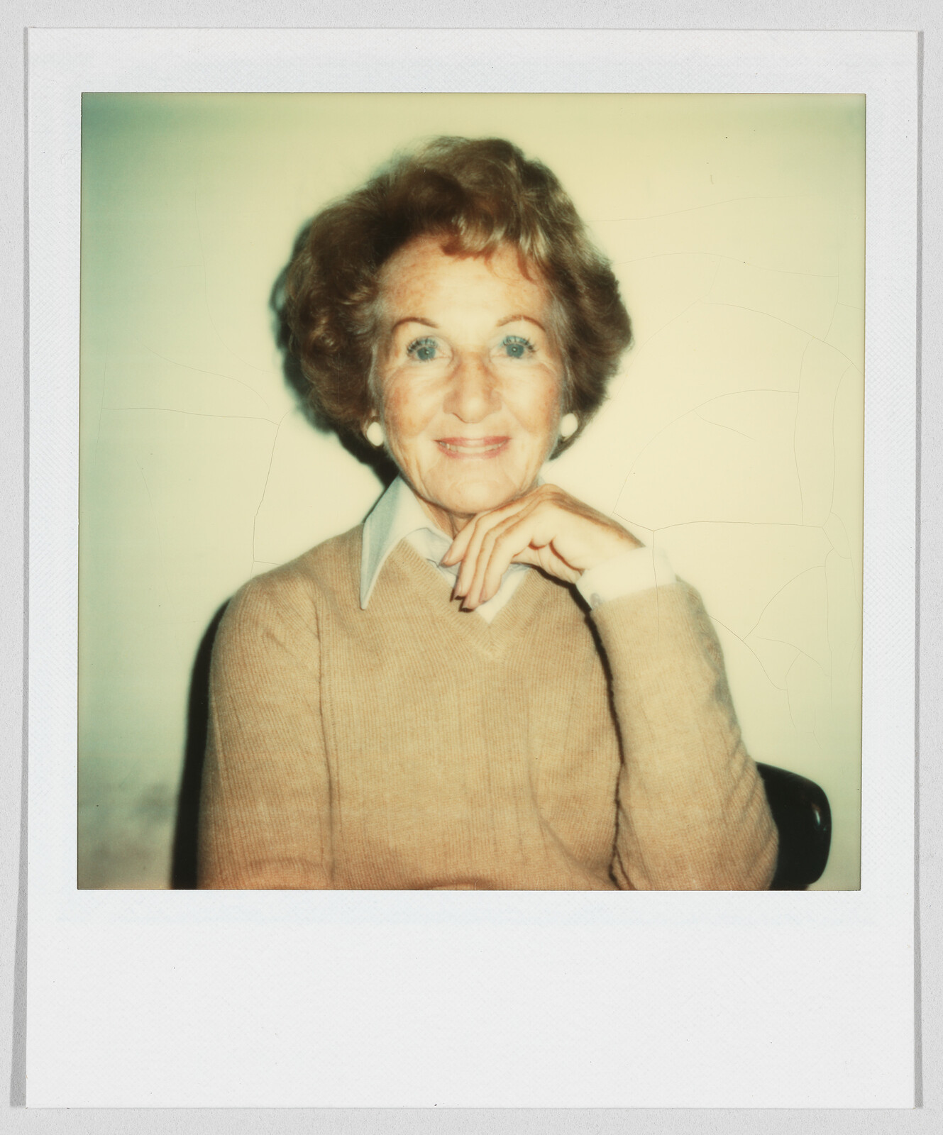 Older woman with short curly hair, wearing a tan sweater and white collared shirt, smiling at the camera.