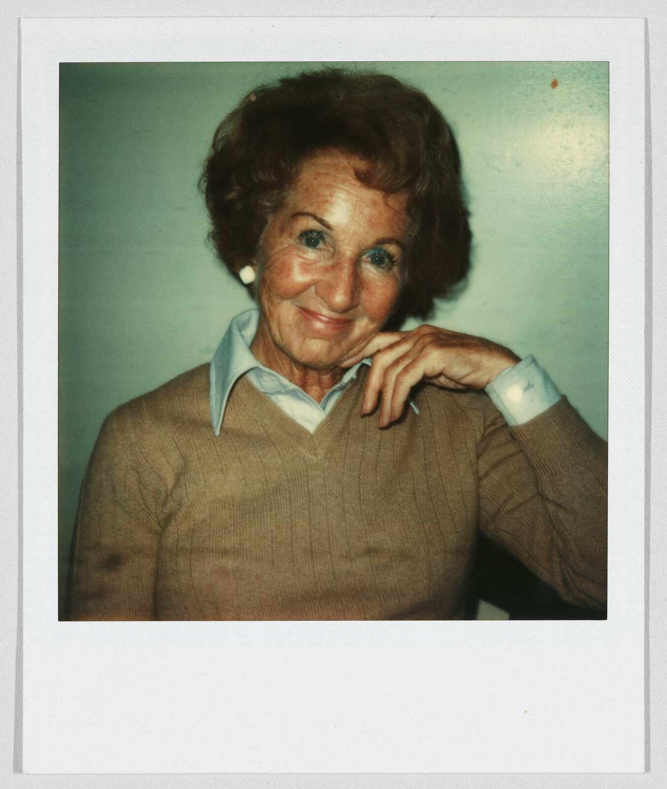 Older woman with short brown hair, wearing a tan sweater and blue collared shirt, smiling at the camera.