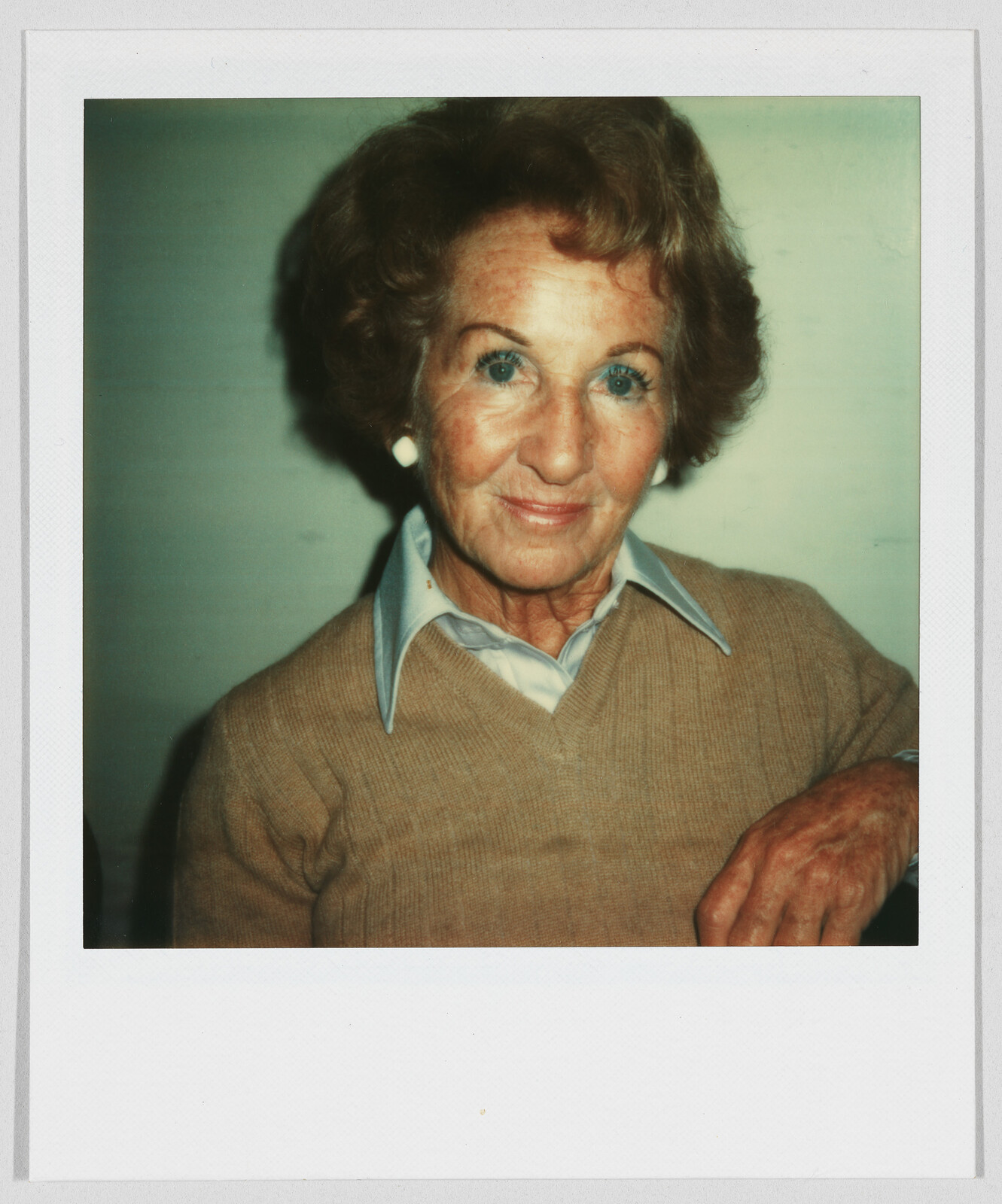 Older woman with short brown hair, wearing a tan sweater and blue collared shirt, smiling gently.