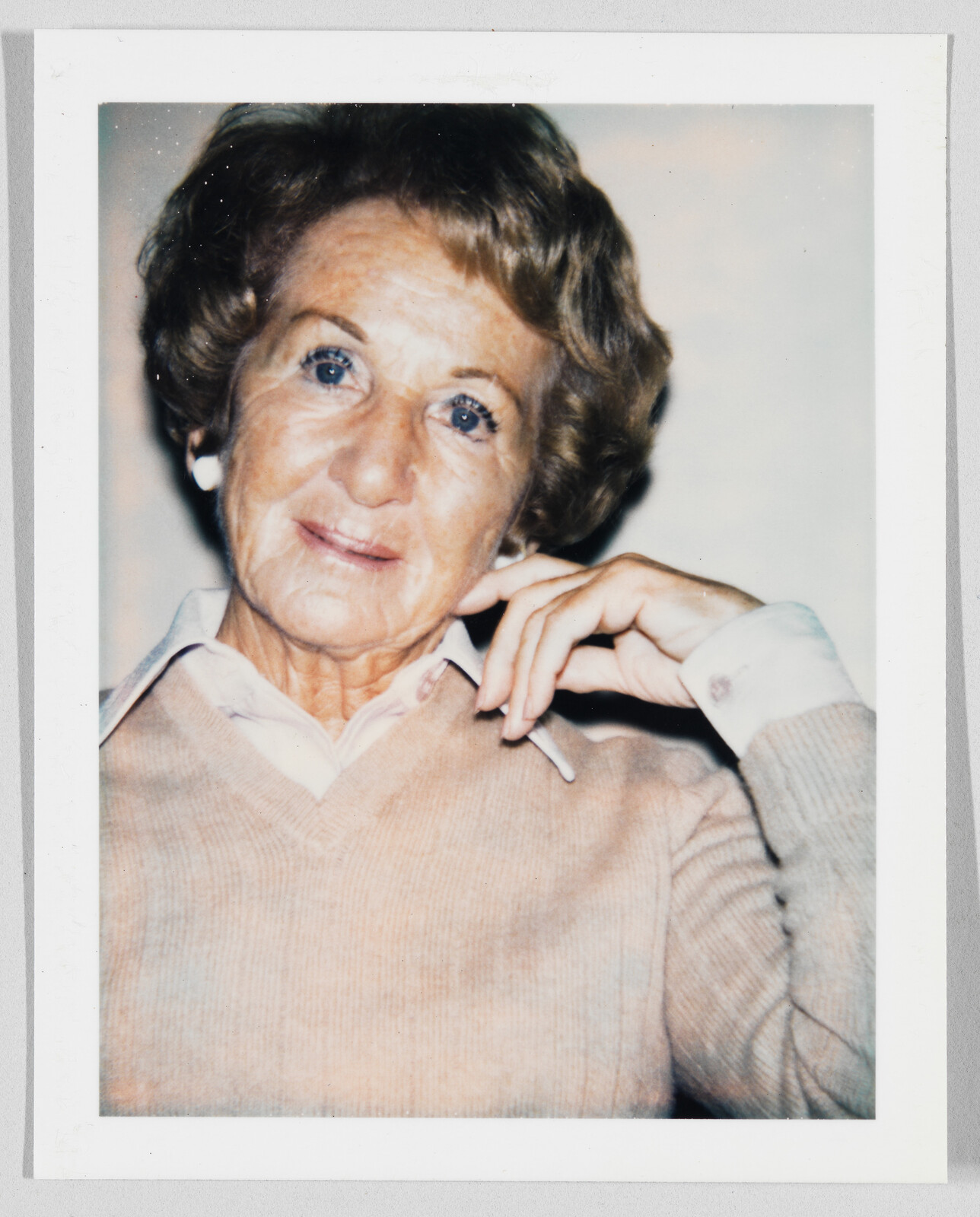Older woman with short hair and light sweater sits, resting her hand near her face, looking at camera.