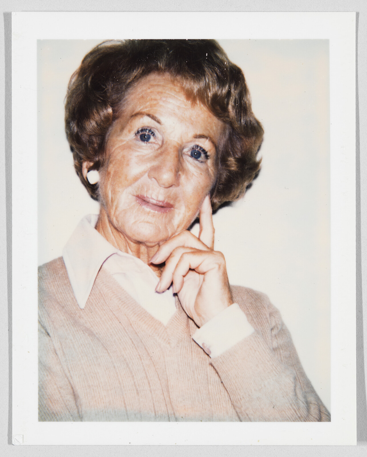 Older woman with short brown hair and white earrings, touching her face and looking at the camera.