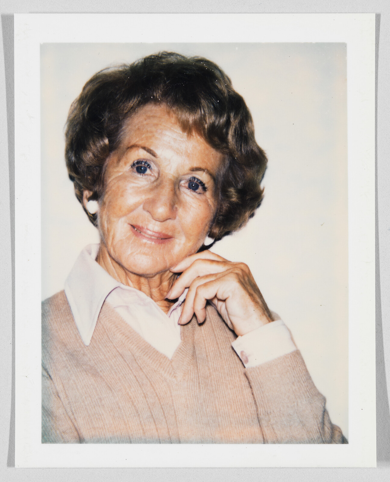 Older woman with short brown hair smiles gently, resting her chin on her hand, wearing a beige sweater.