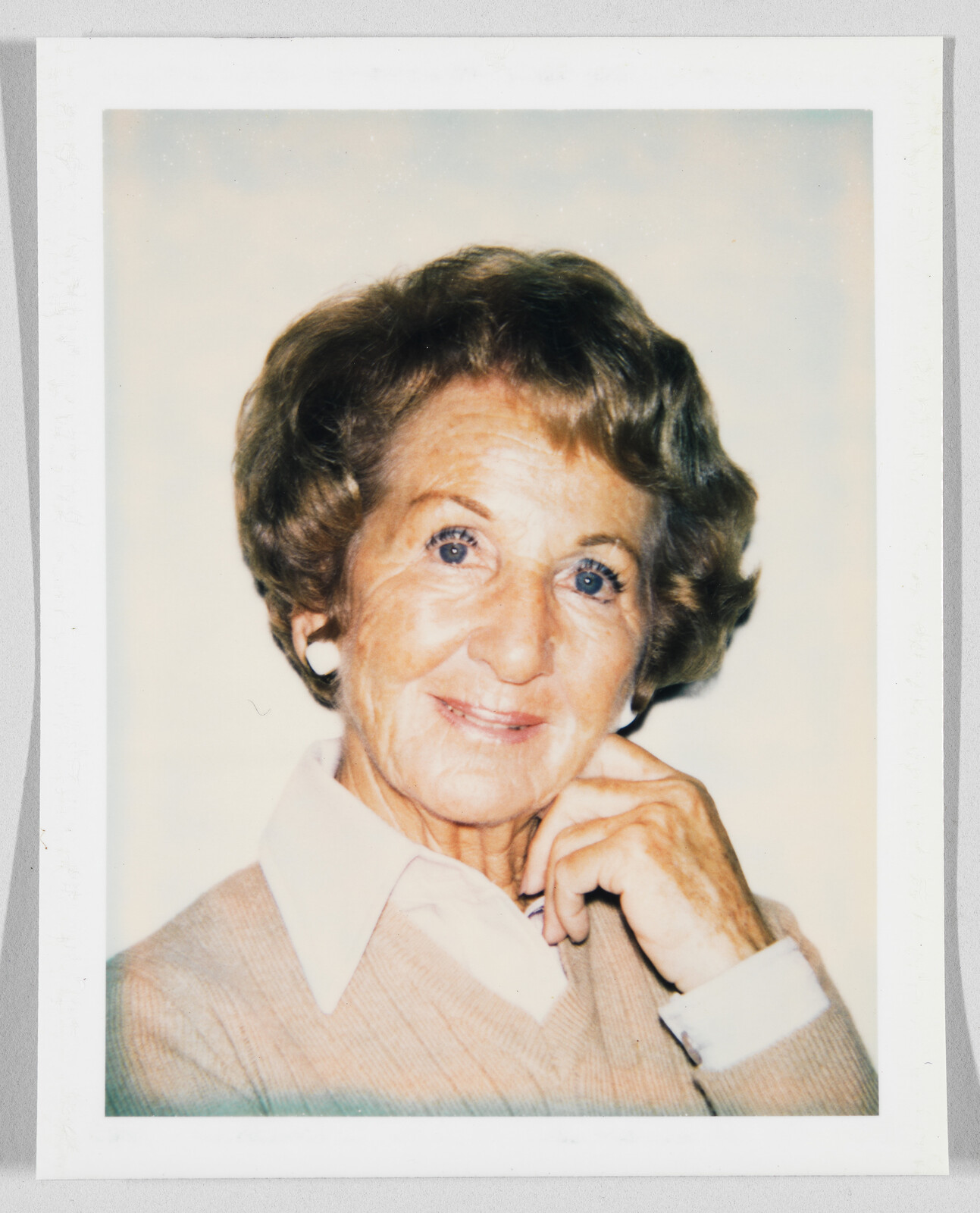 Older woman with short brown hair, wearing a beige sweater and white collared shirt, smiling gently.