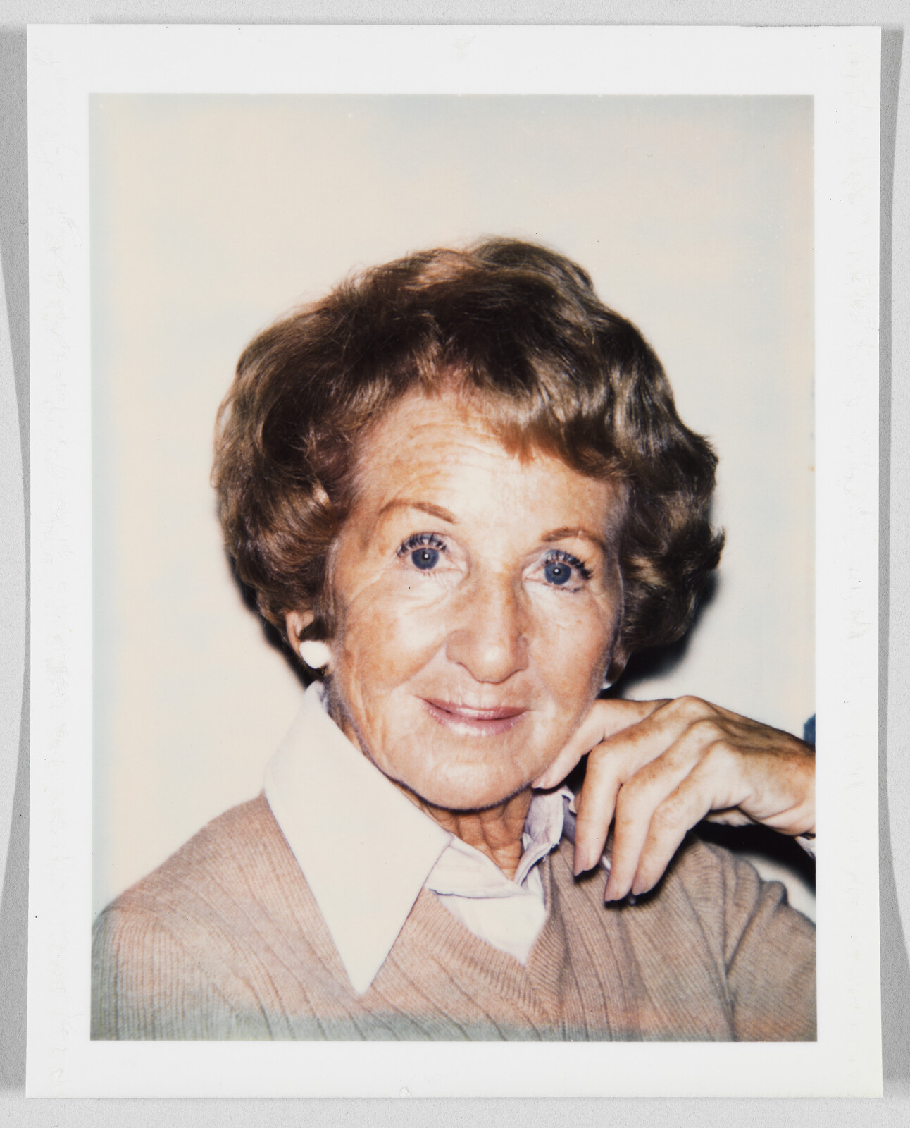 Older woman with short brown hair, wearing a light sweater and collared shirt, smiling and resting her chin on her hand.