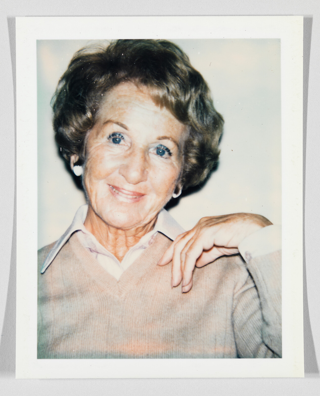 An older woman with short hair smiles gently, wearing a light sweater and collared shirt, hand resting near chin.