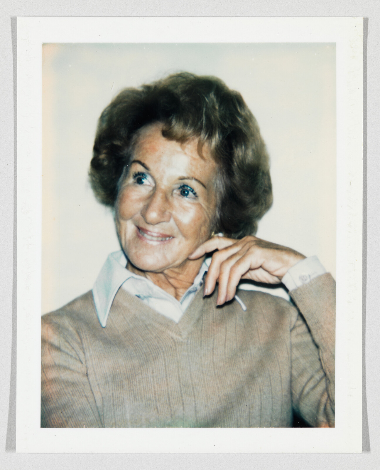 An older woman smiles and looks to the side with her hand resting near her face.