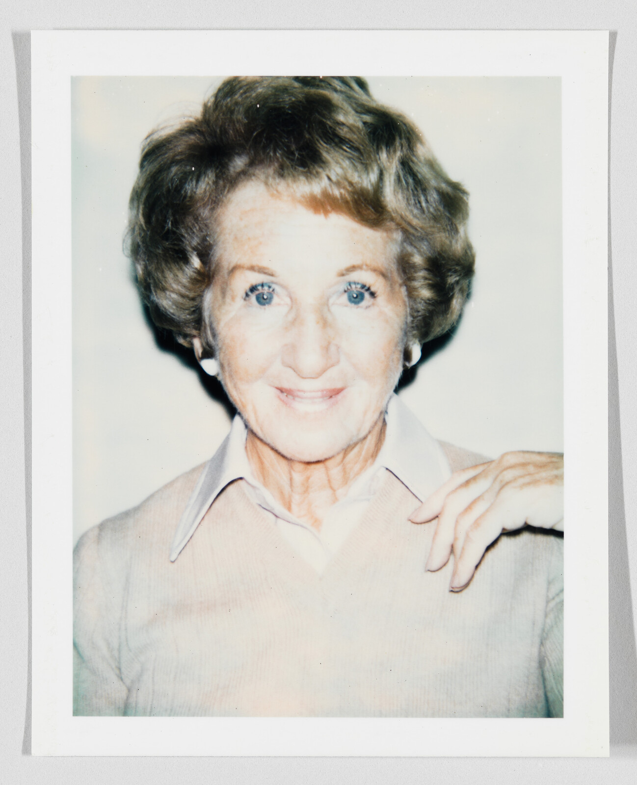 Elderly woman with short hair smiling at camera, hand resting on her shoulder.