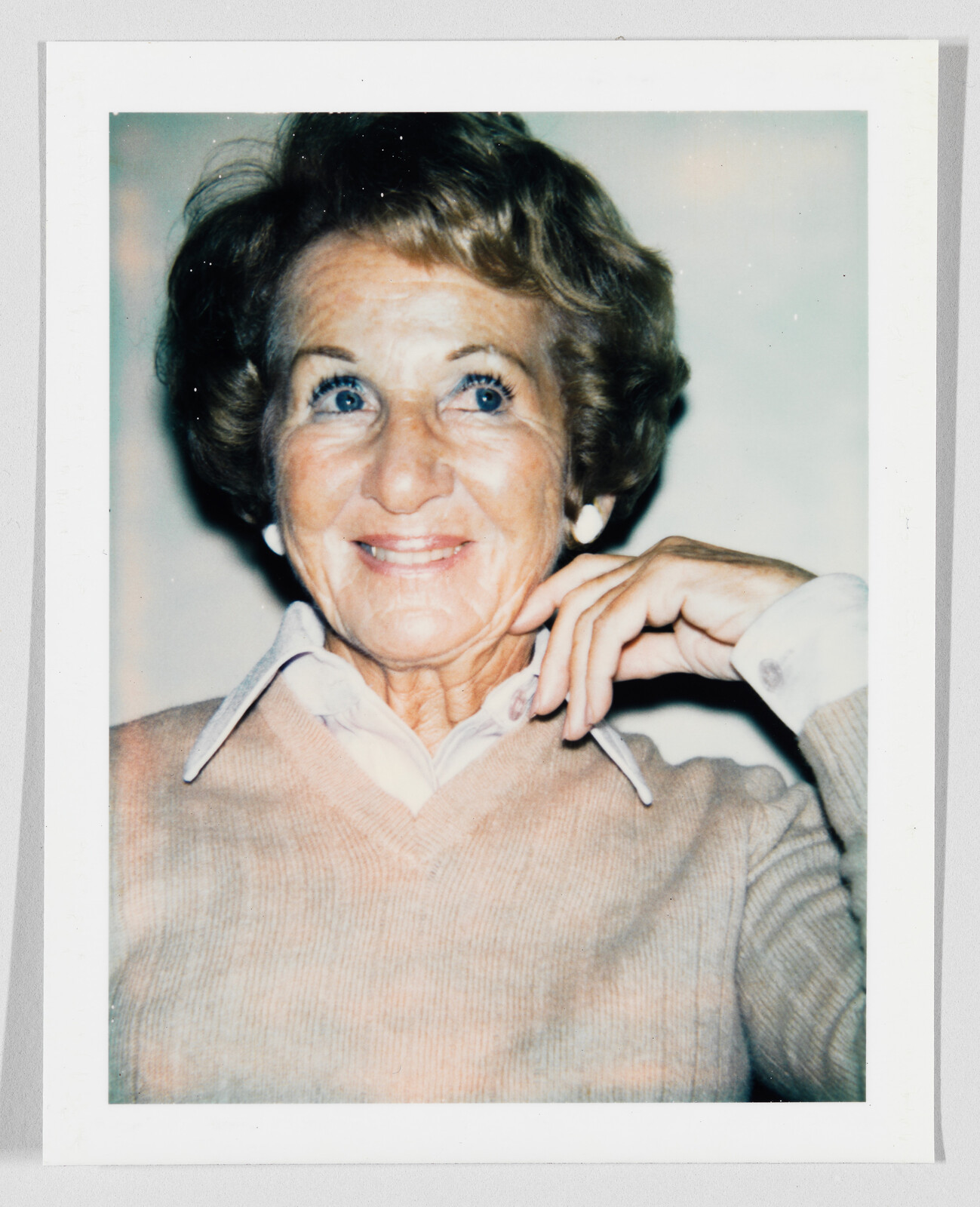 An elderly woman smiles while resting her hand against her cheek and looking upward.