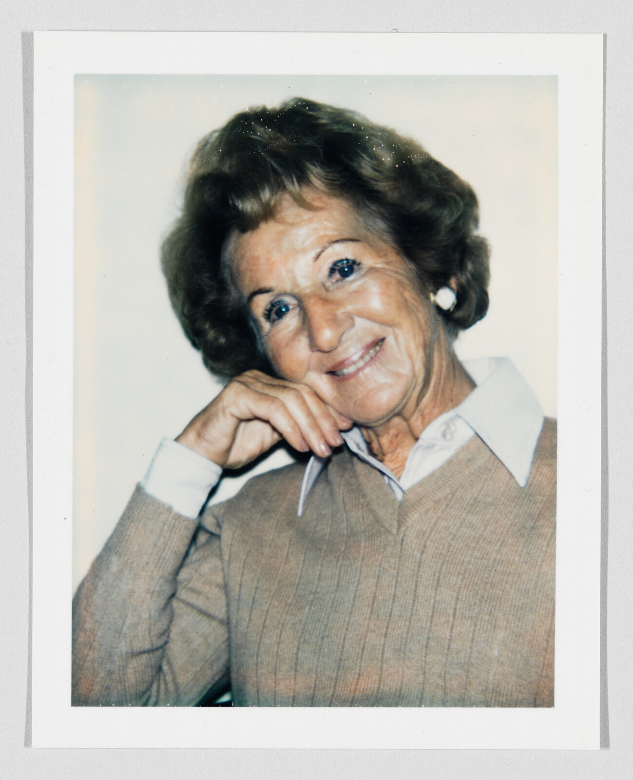 Older woman with short brown hair smiles, resting her chin on her hand, wearing a tan sweater and collared shirt.