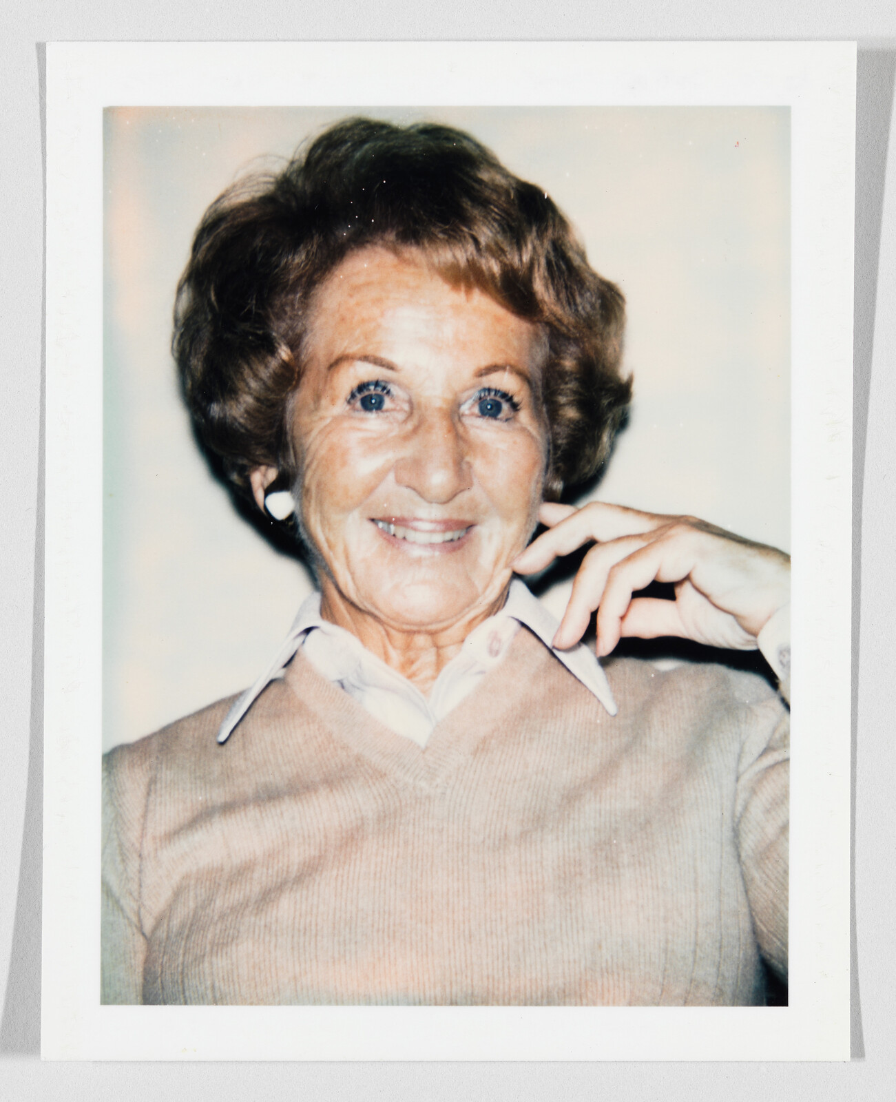An elderly woman smiles at the camera with her hand resting near her cheek.