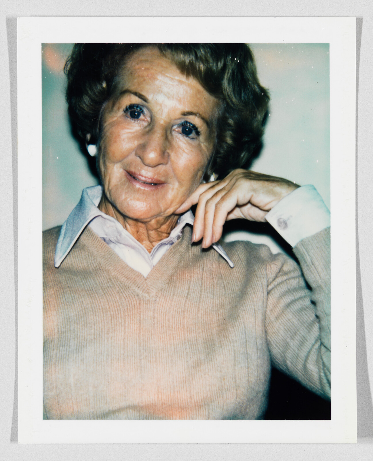 An elderly woman smiles gently with her hand resting near her face and collar.