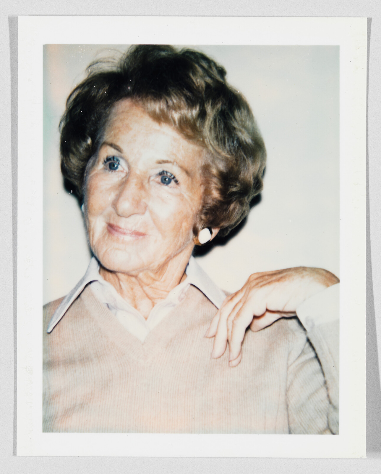 An elderly woman with white earrings smiles softly while resting her hand on her shoulder.