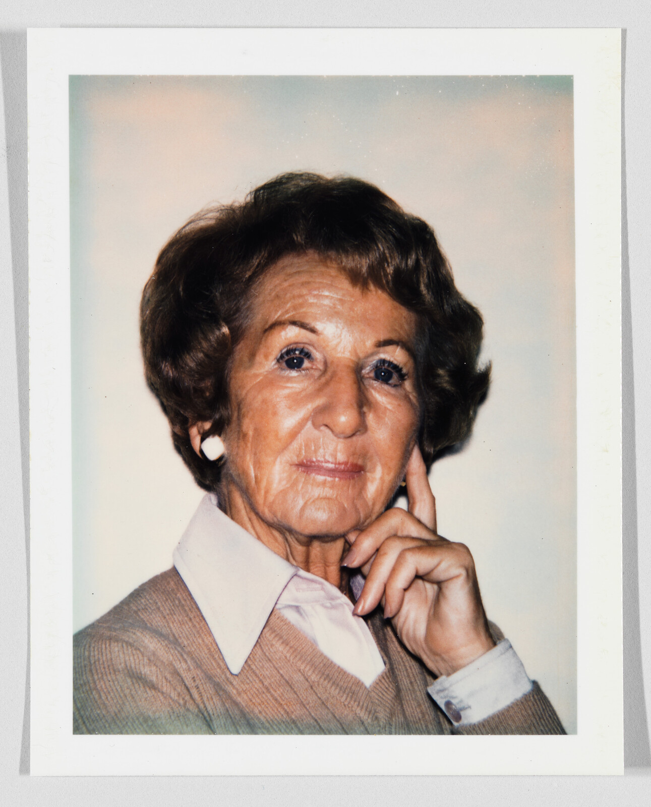 Elderly woman poses thoughtfully with her hand resting on her cheek, wearing a collared shirt.