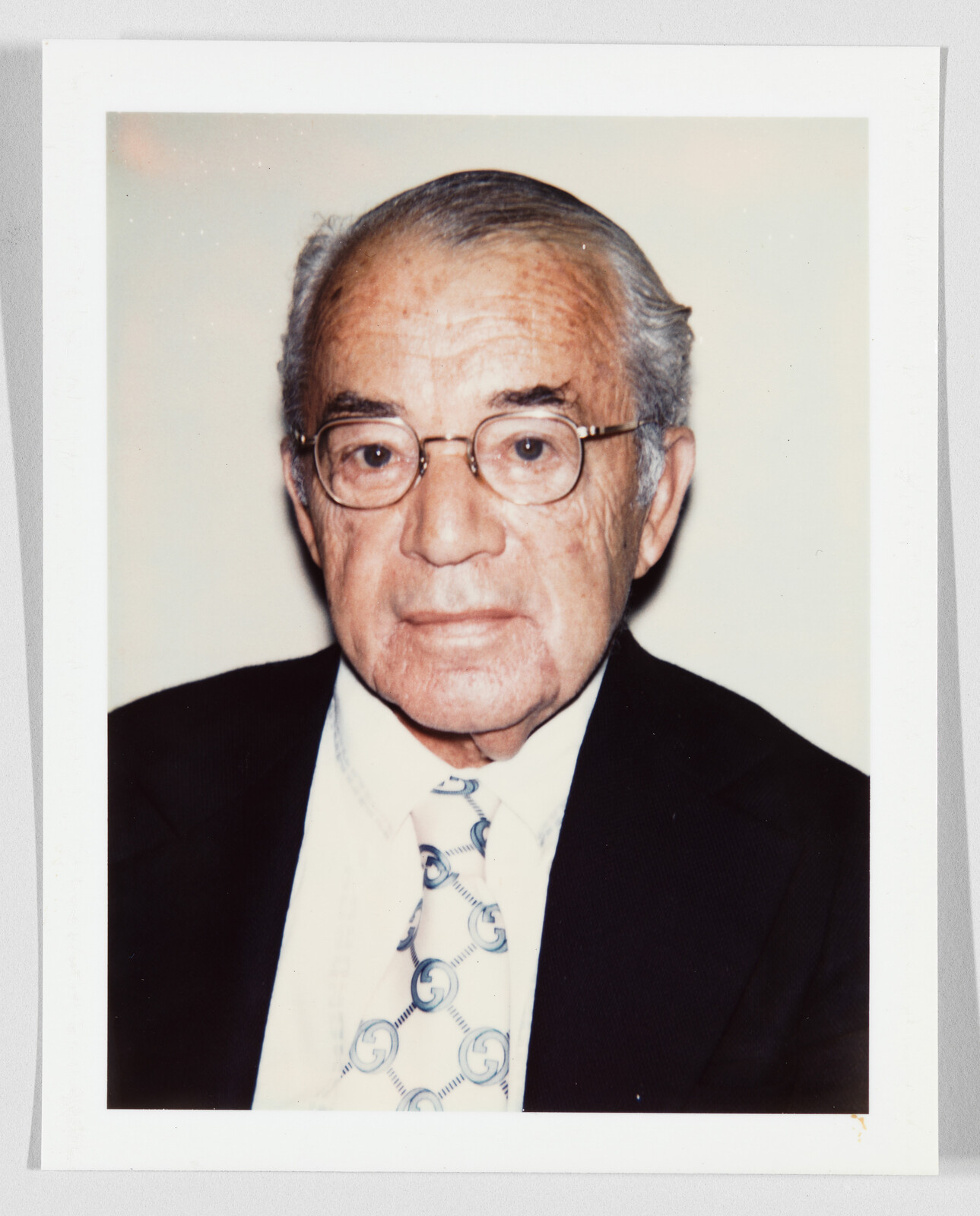 Elderly man with glasses and patterned tie wearing a dark suit and neutral expression.