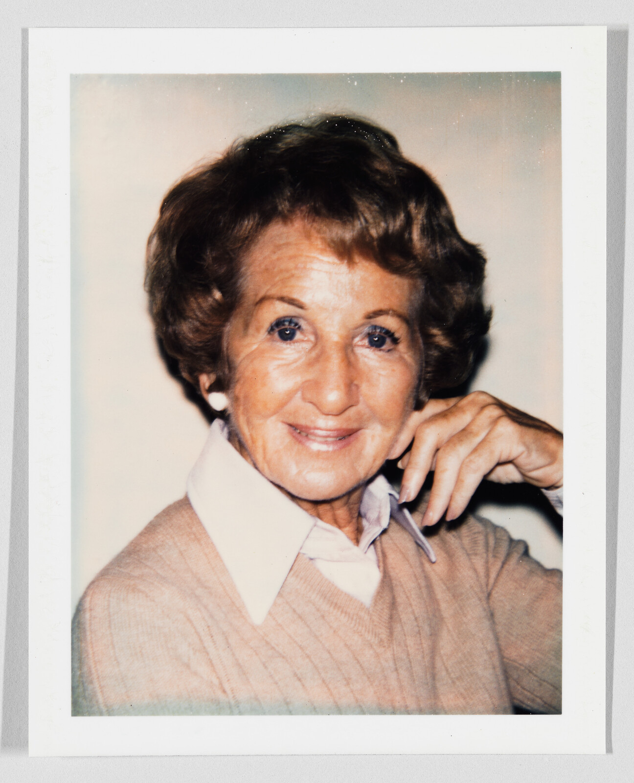 An older woman smiles with her hand resting near her chin, wearing a collared sweater.