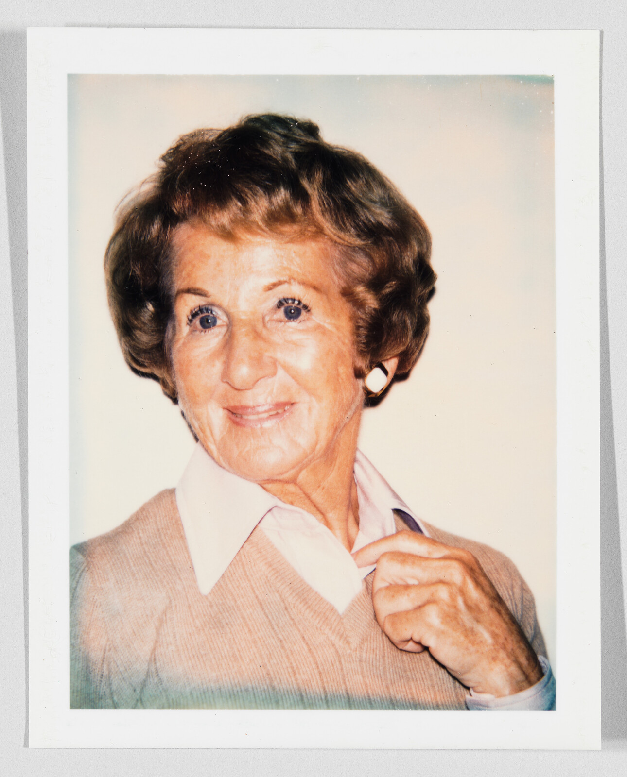 An older woman smiles while adjusting her shirt collar, wearing a beige sweater and white earrings.