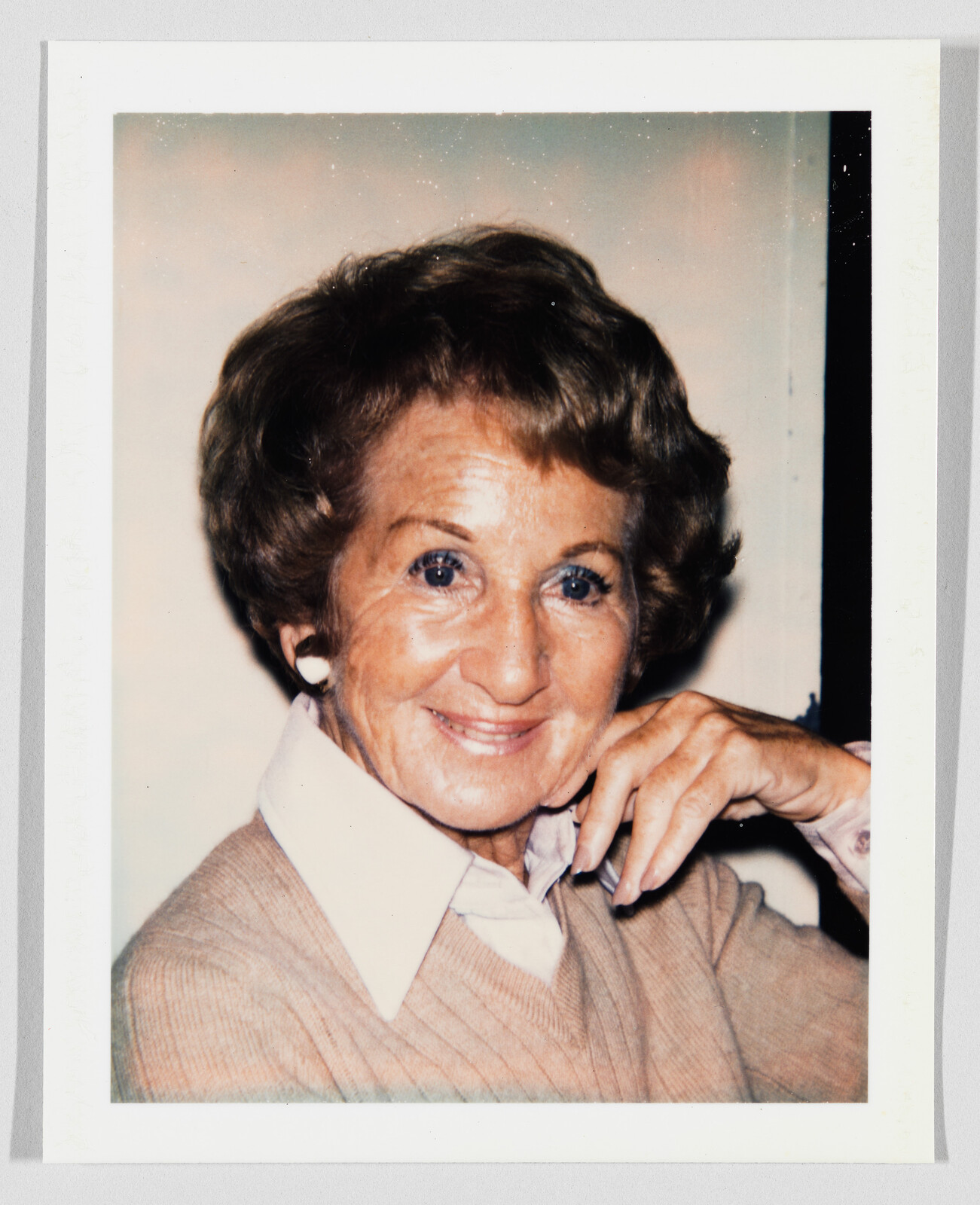 Elderly woman smiling gently while resting her chin on her hand and wearing a sweater.
